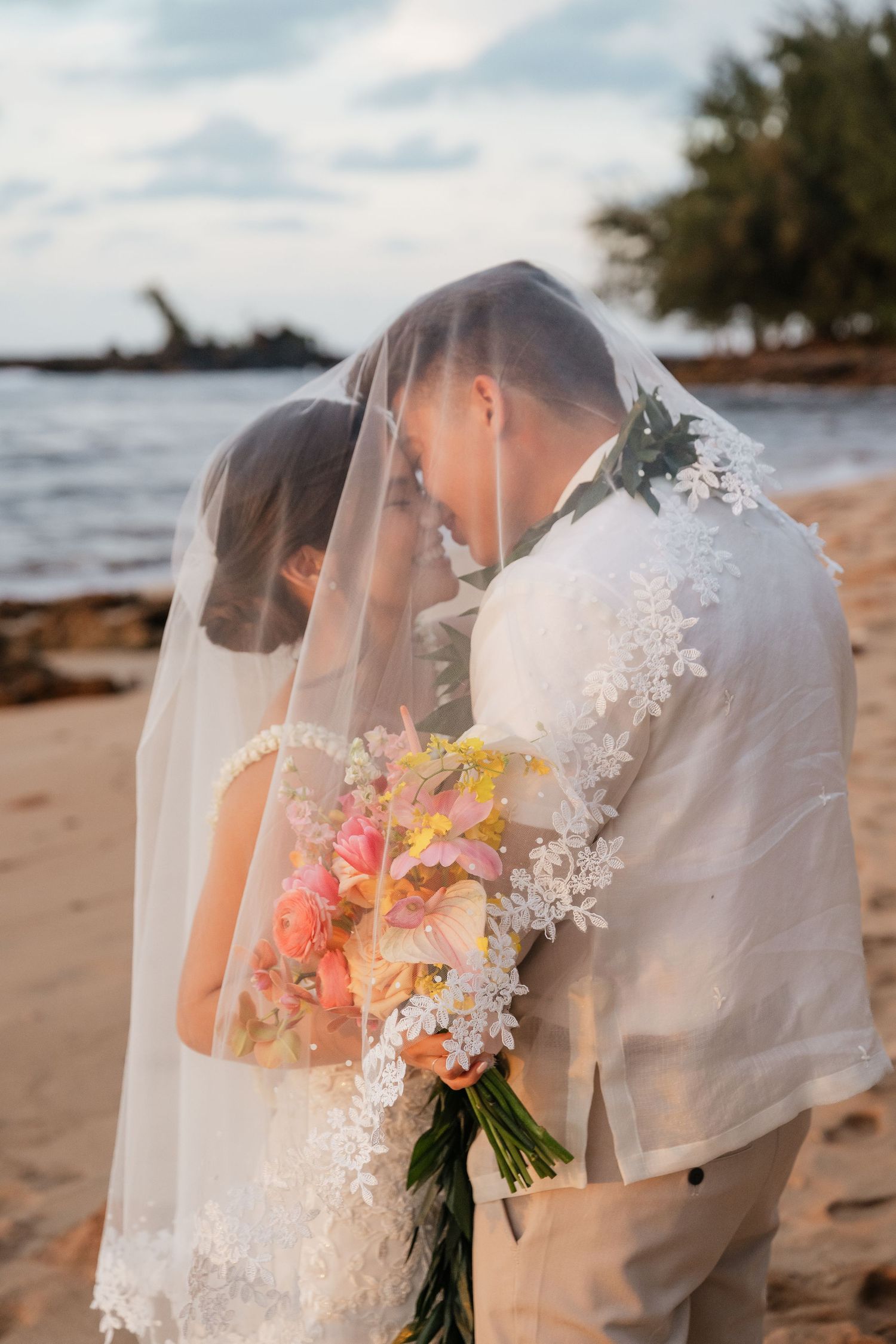 Bride and groom sharing a romantic moment under a lace veil on a sandy beach, holding a colorful tropical bouquet.
