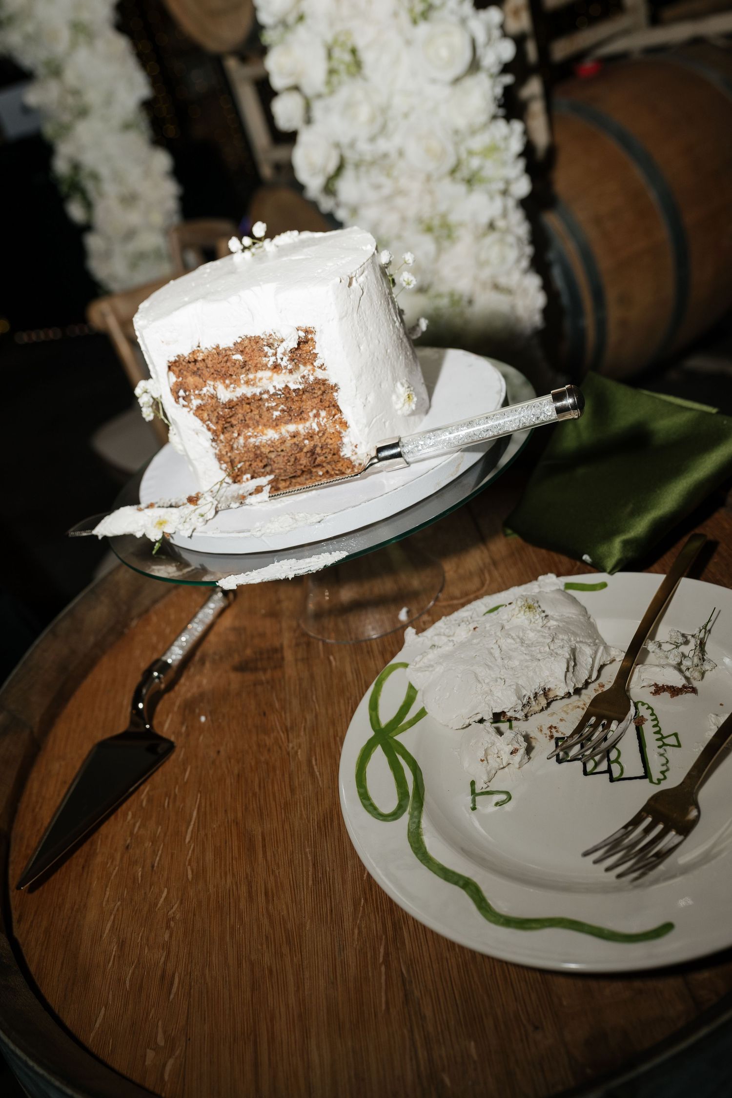 White frosted wedding cake with a server on a rustic wooden barrel beside a dessert plate.