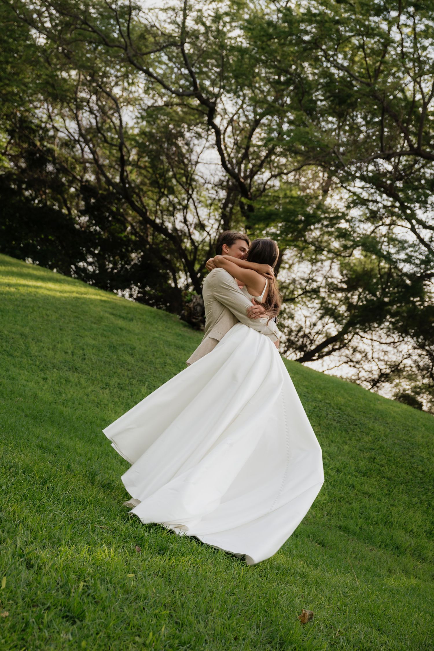Wedding couple embraces on a grassy hillside with trees in the background during golden hour.