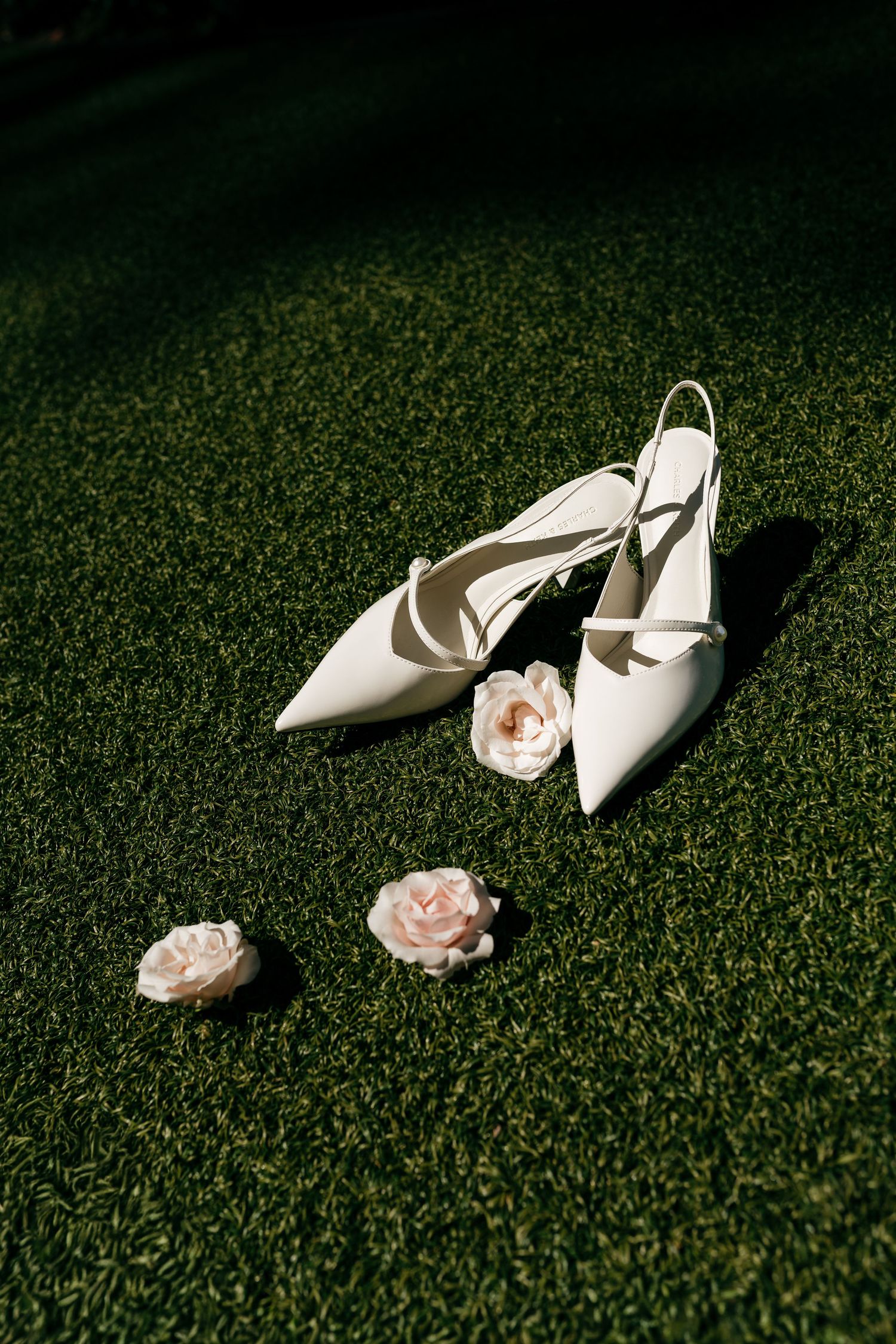 White slingback bridal heels placed on green grass alongside delicate pink roses in natural sunlight.