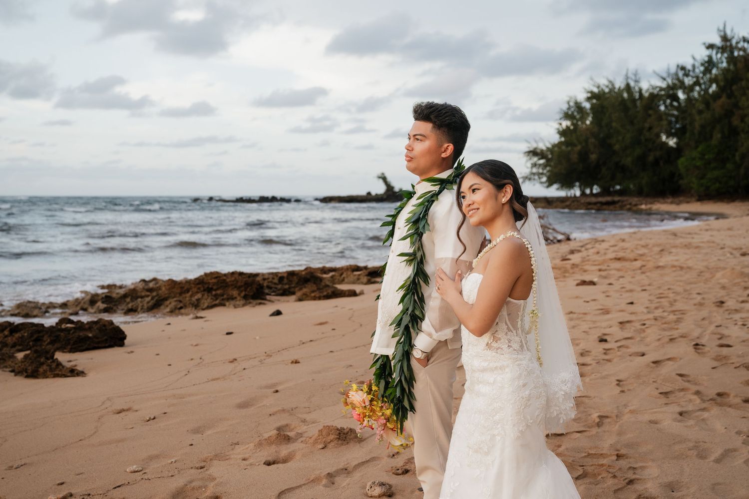 Couple in wedding attire walks on Hawaiian beach, groom wearing lei, bride holding bouquet in white gown with veil.