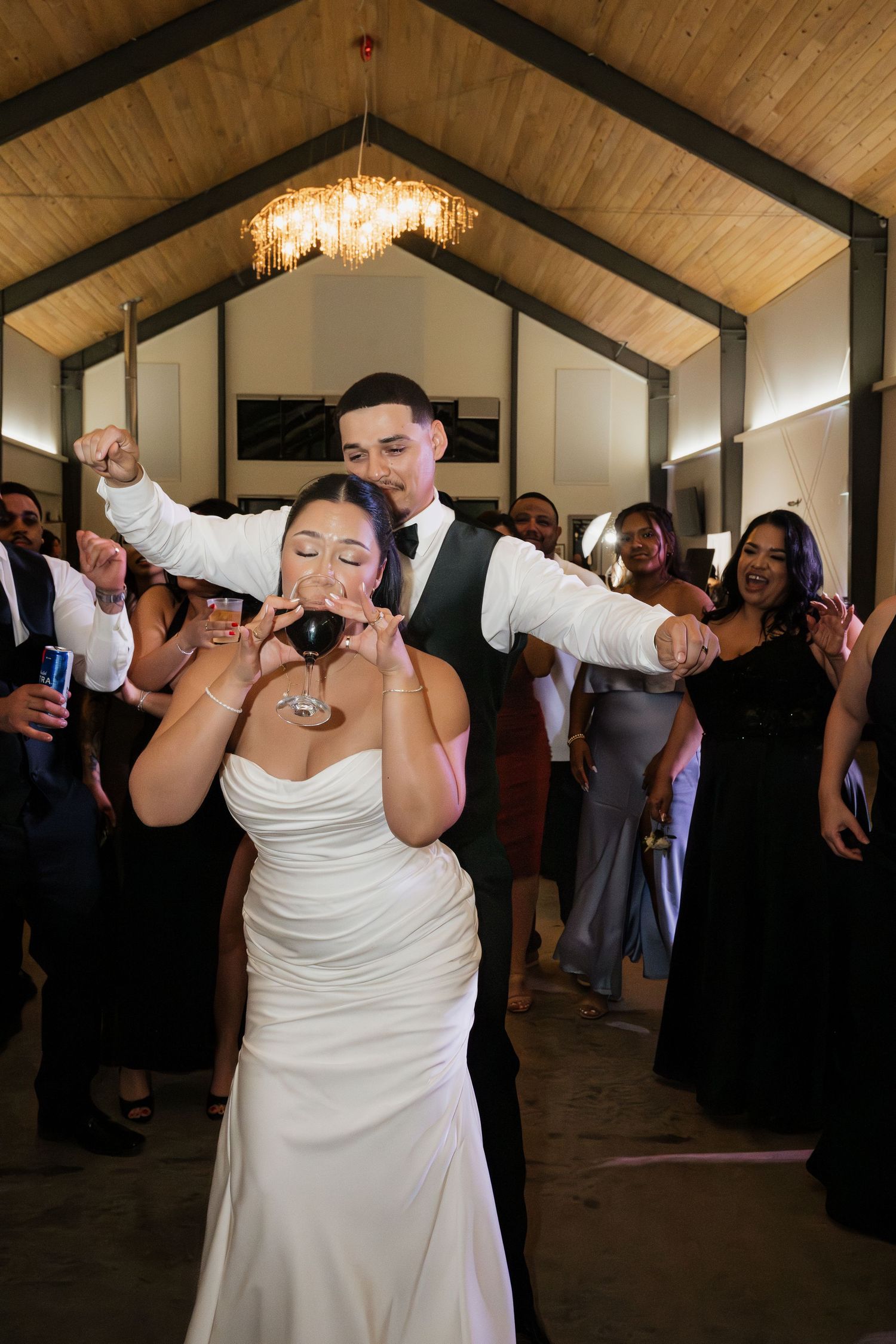 Bride in white gown drinks red wine while groom celebrates behind her at a lively wedding reception.