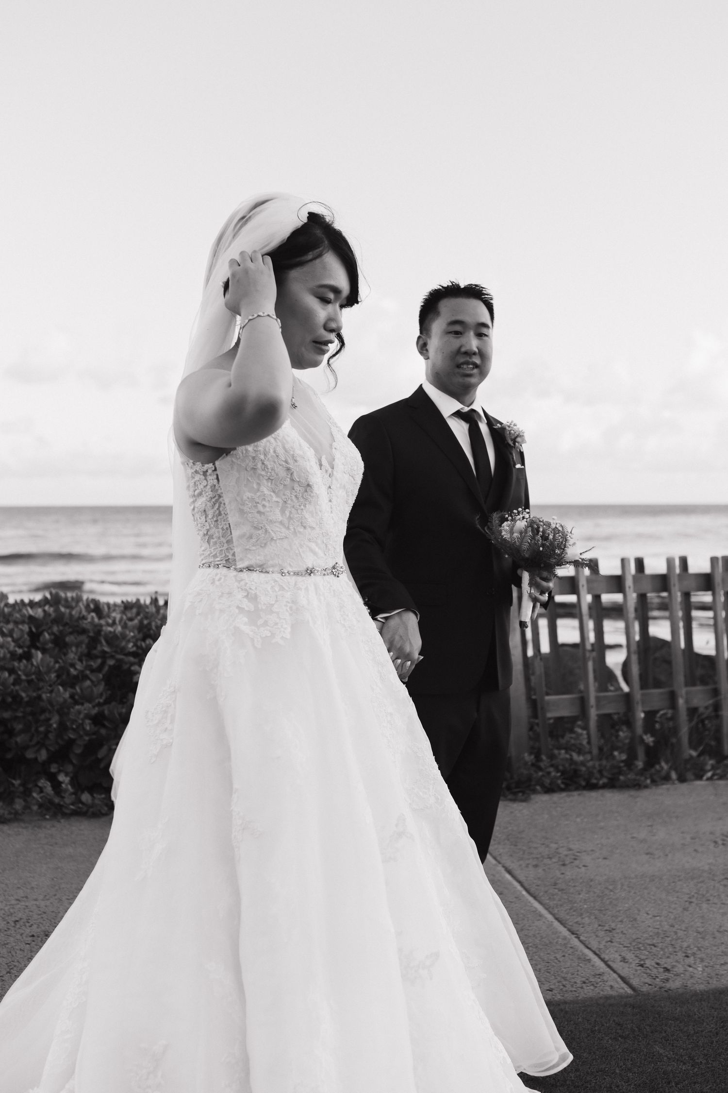 A couple in wedding attire walking together on a beachside path in black and white.