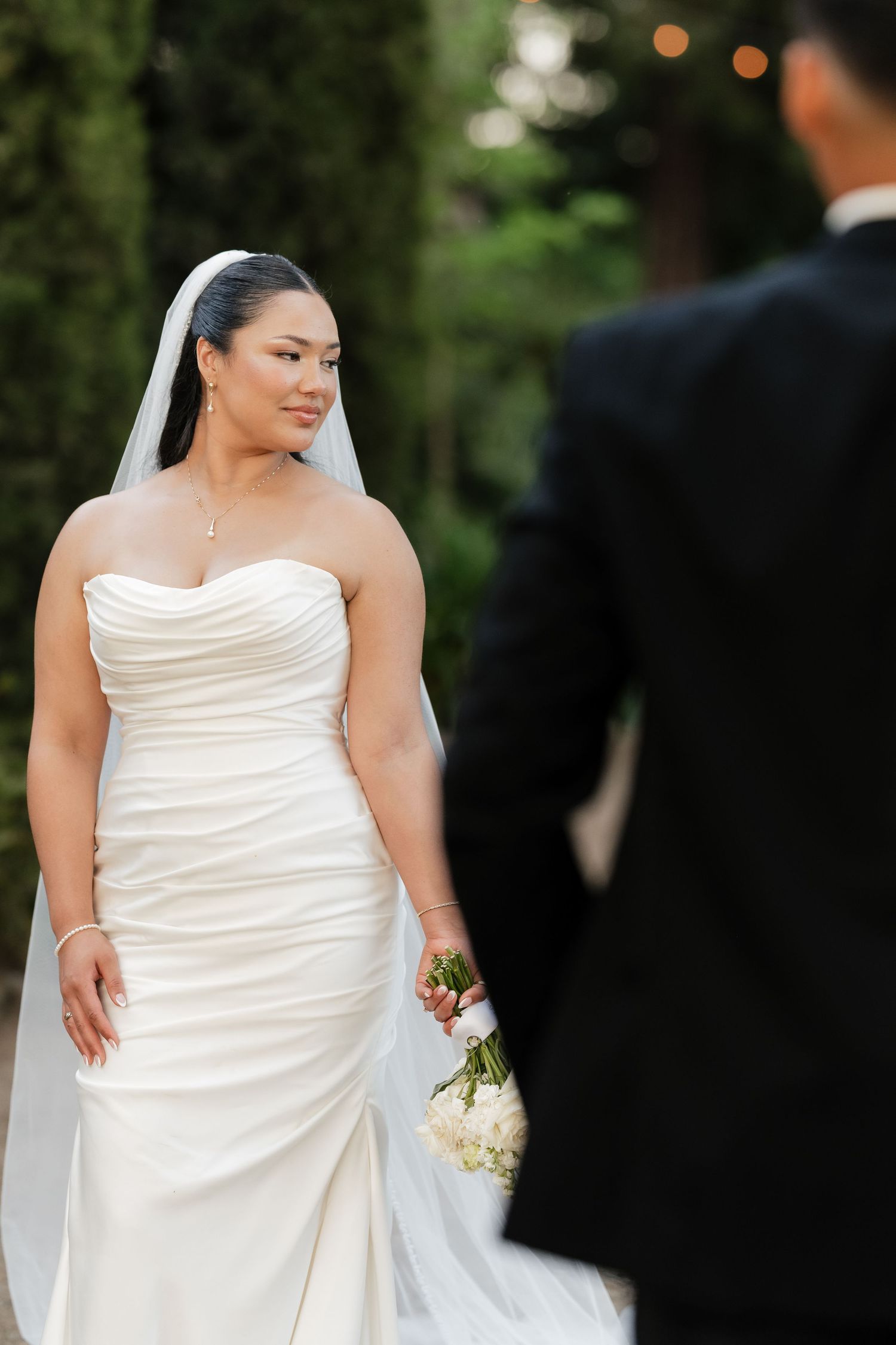 Bride in strapless white gown and veil holds bouquet, smiling at groom in black suit outdoors.
