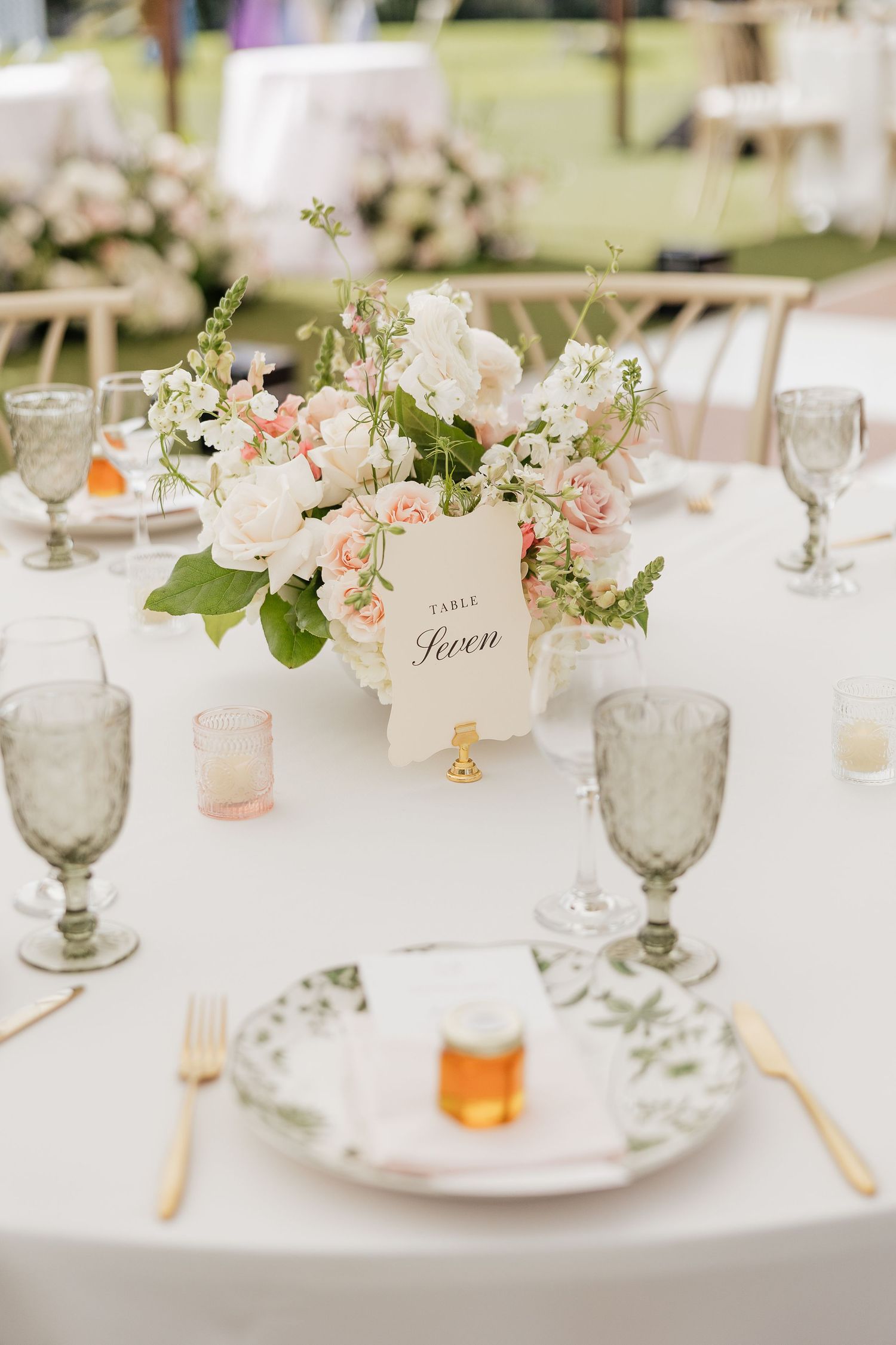 Elegant wedding reception table seven with white floral centerpiece, green goblets, botanical plates, and gold cutlery.