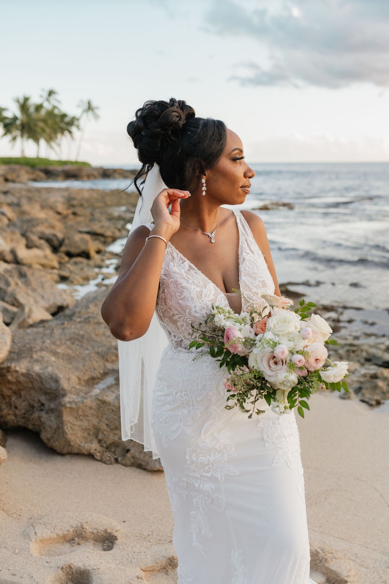 Bride in lace dress holds a white bouquet against rocky beach landscape.