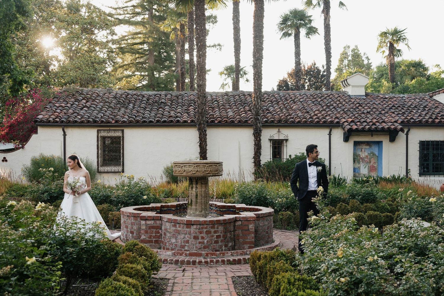 Bride and groom stand by a brick fountain in a lush garden beside a white Spanish-style villa with palm trees.