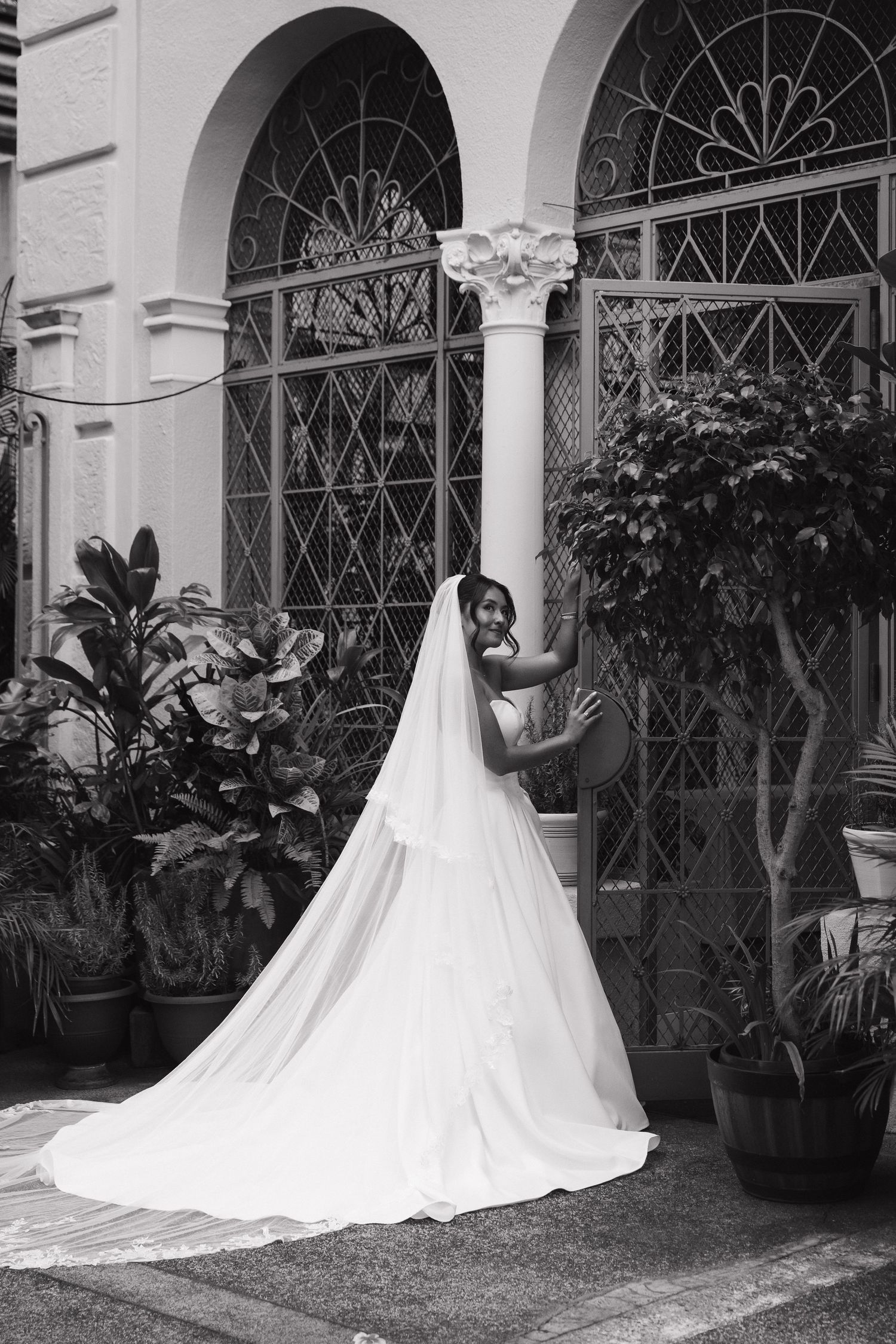 Black and white photo of a bride in a flowing wedding gown and veil posing by ornate arched windows with lush plants.