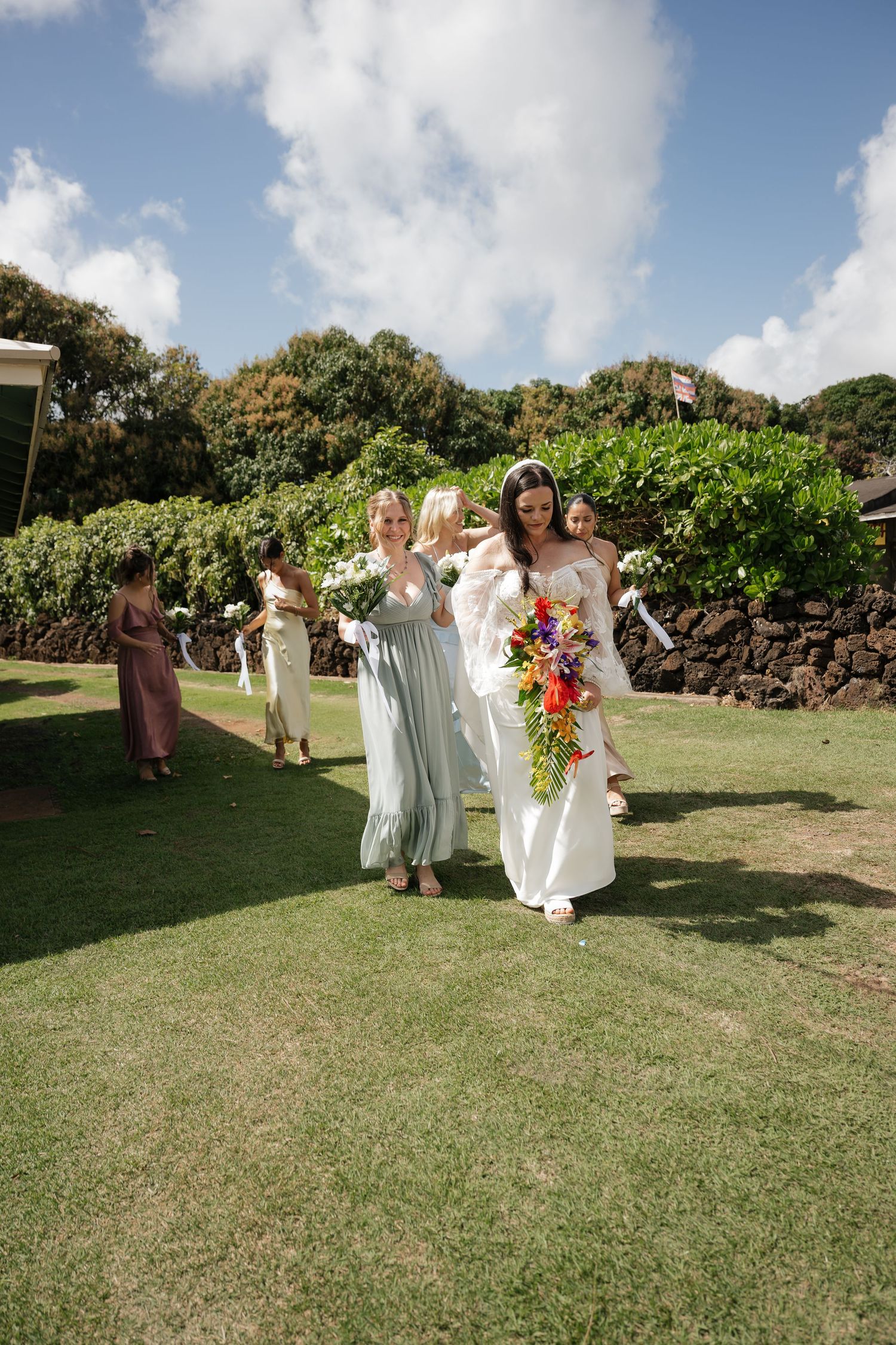 Wedding party walks across sunny lawn with tropical landscaping and stone wall.