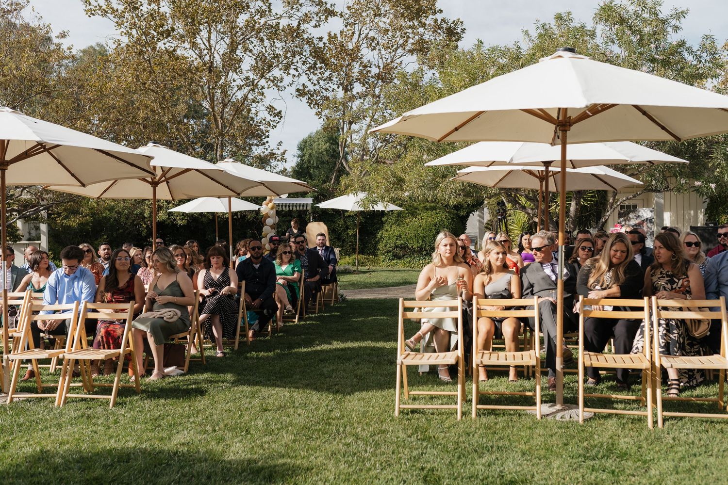 Outdoor wedding ceremony setup with wooden benches and white umbrellas in a garden setting.
