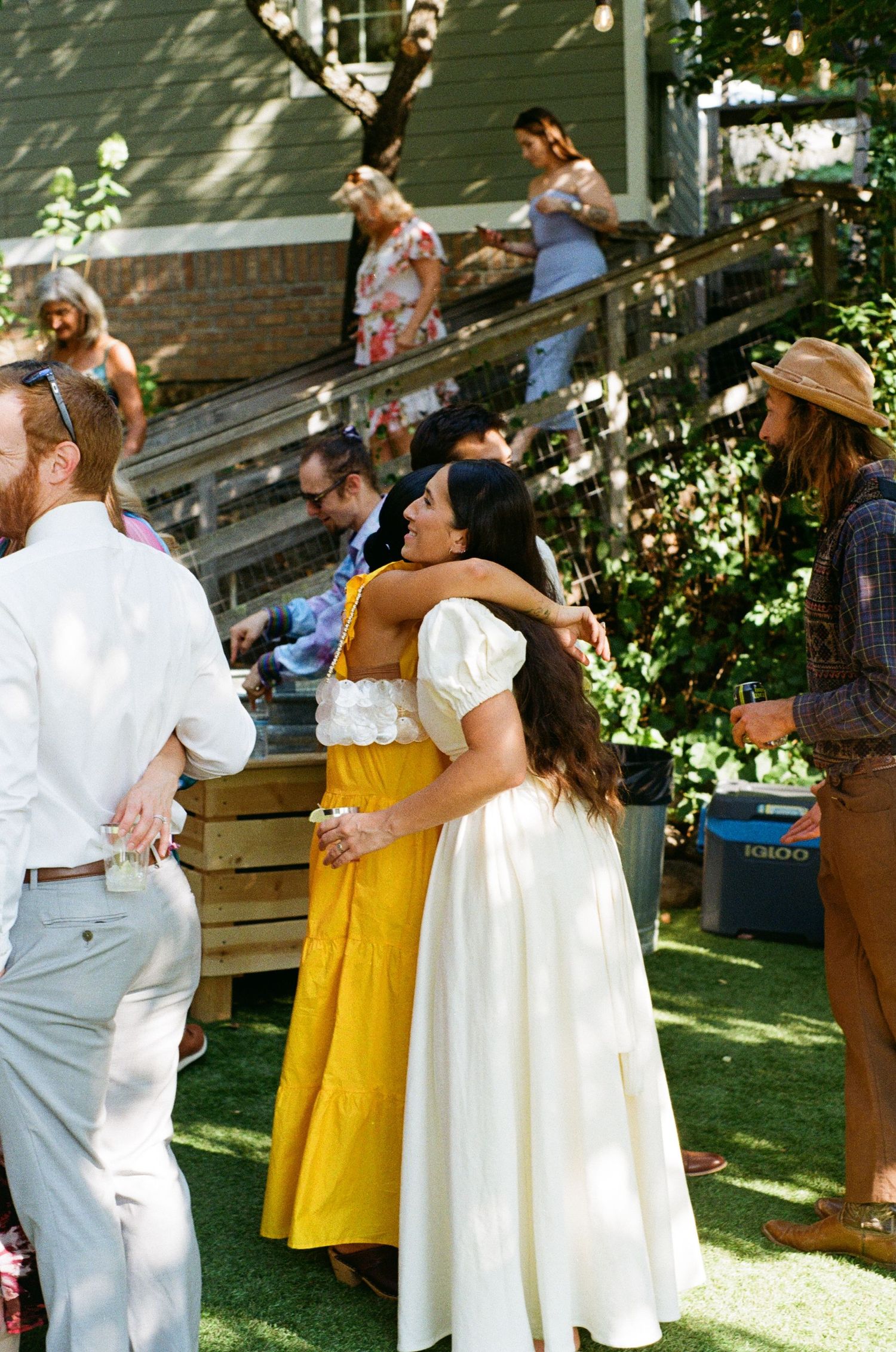 Wedding guests dance and embrace during outdoor garden reception on sunny afternoon.