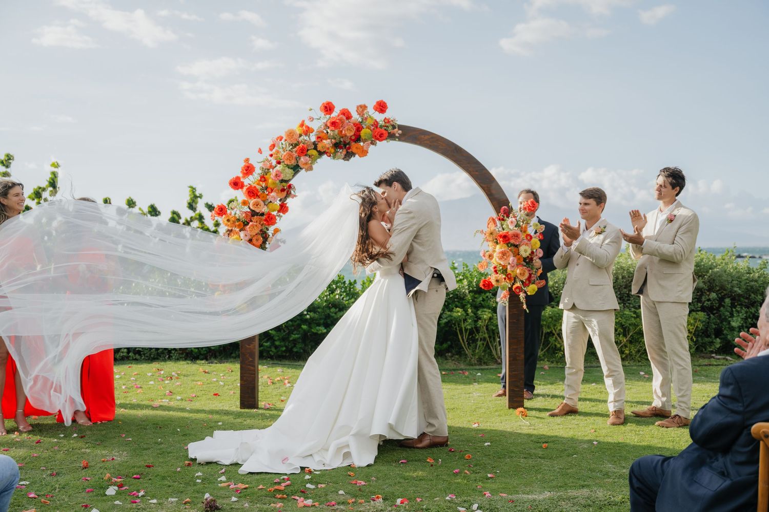 A wedding party poses on a grassy hilltop with flowing white dress and orange floral arrangements in the breeze.