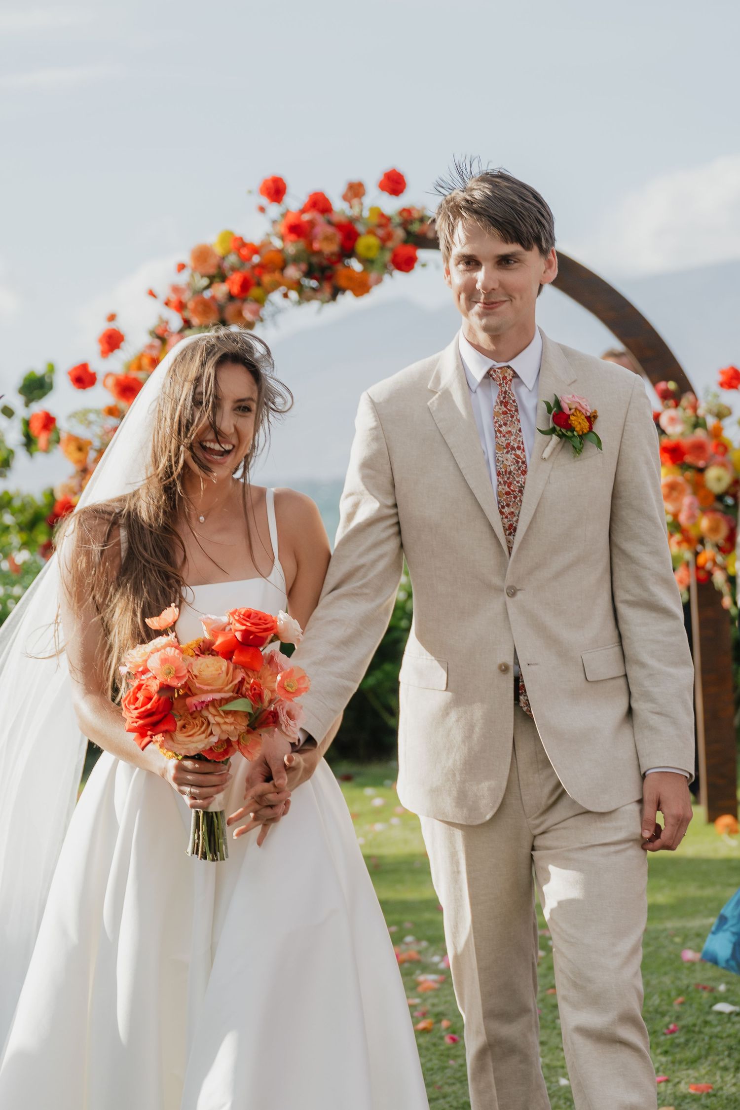 Wedding party in tan suits and white dress poses with vibrant coral flower arrangements.