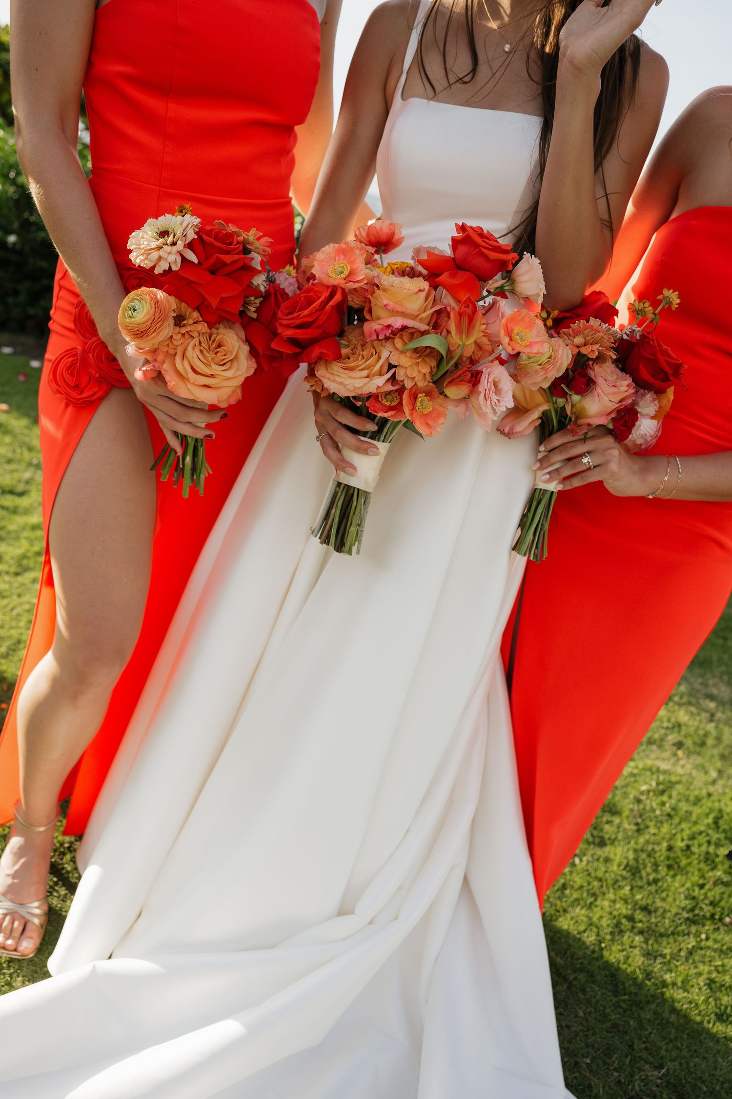Bridesmaids in coral red dresses hold matching floral bouquets against a white wedding dress.