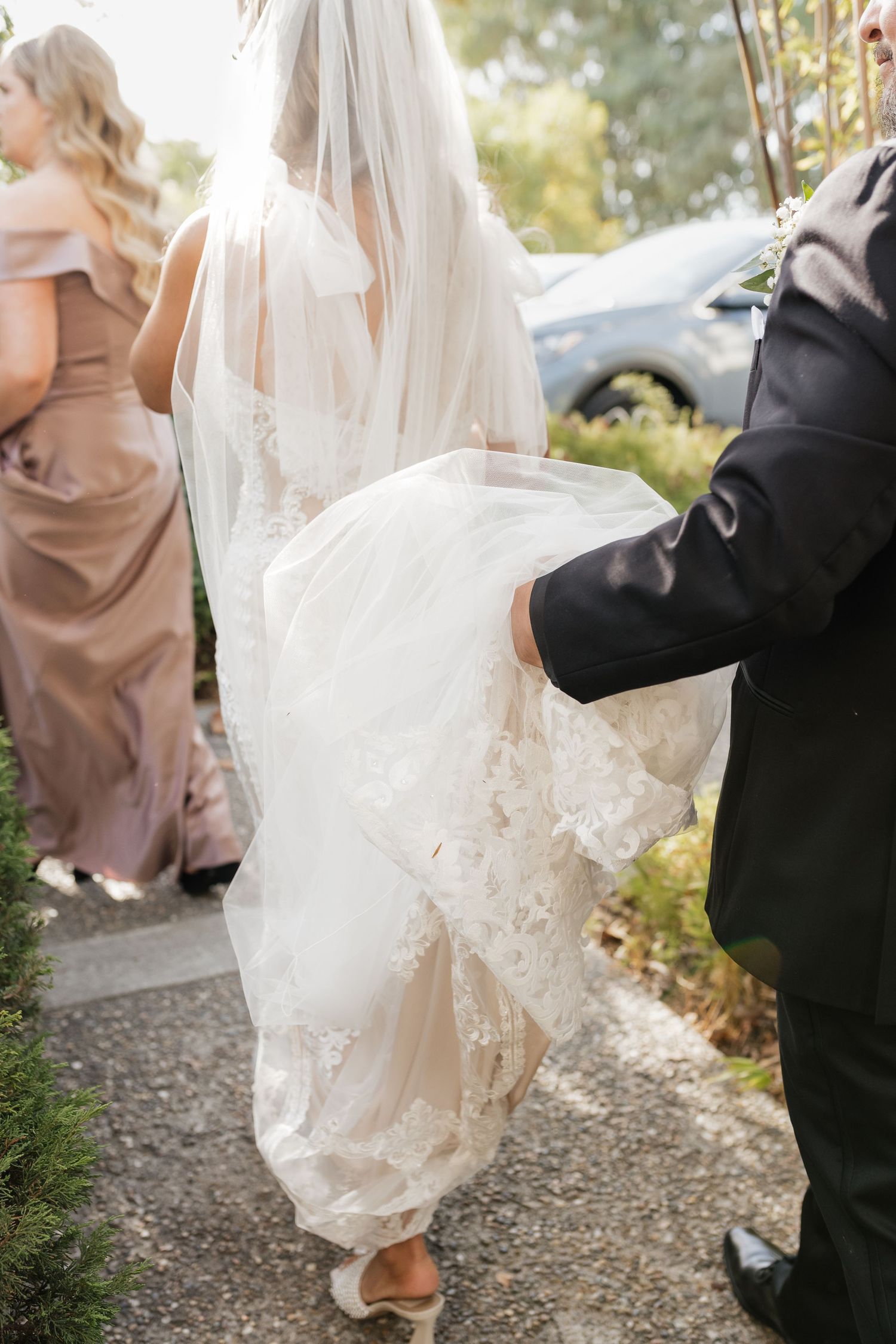 Detail shot of lace wedding dress and veil being carried while walking on garden path.