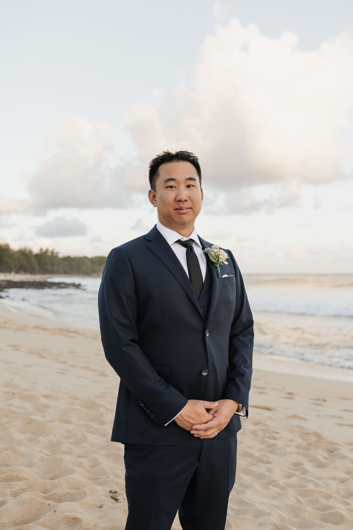 A groom in a navy suit standing on a beach with cloudy sky backdrop.