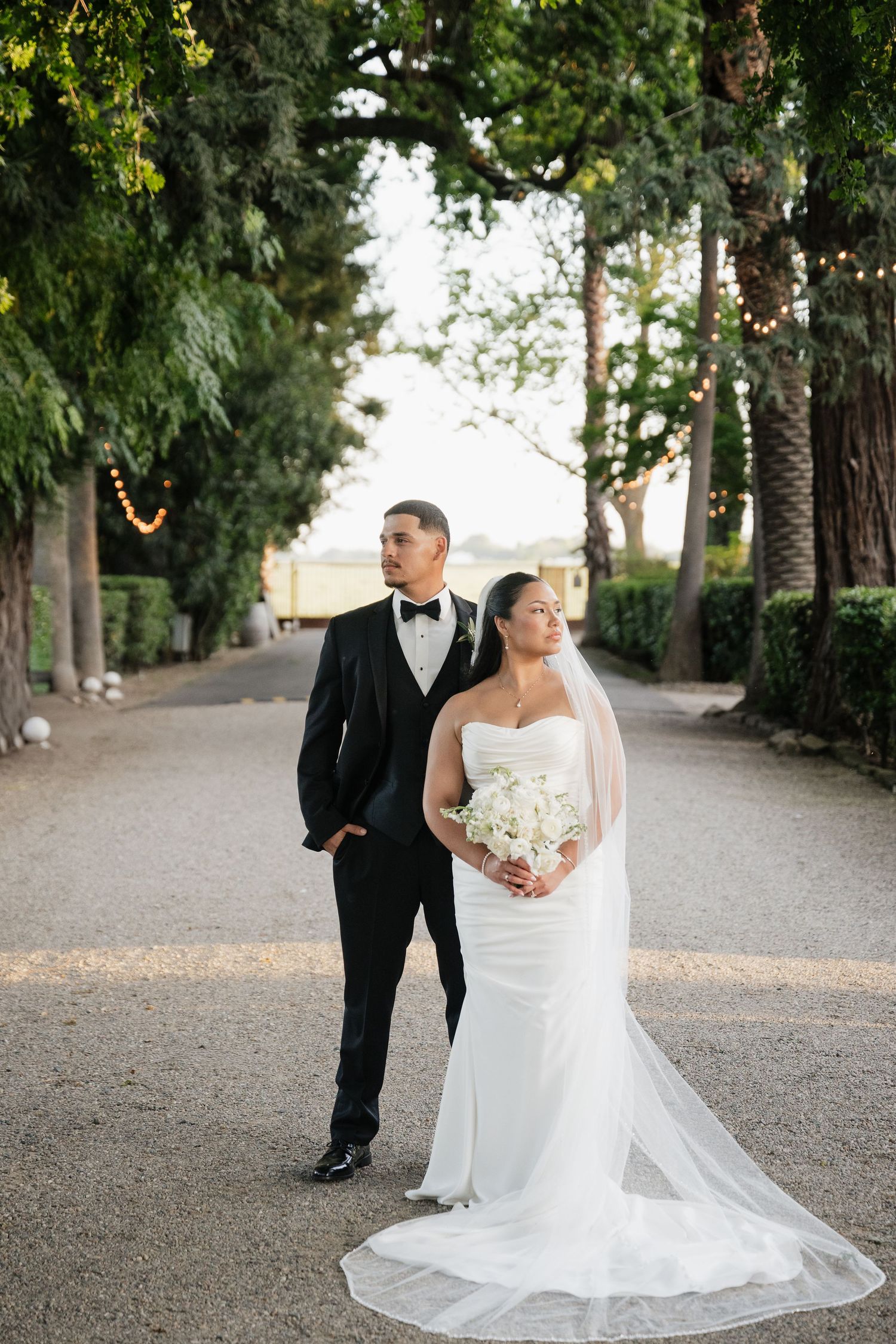 Bride and groom posing on a tree-lined gravel path, she in a strapless gown with veil, he in a black tuxedo.