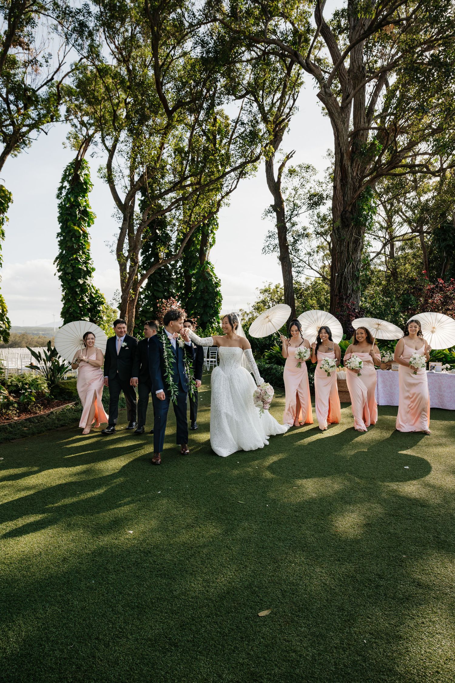 Wedding party walks on green lawn, bridesmaids in pink dresses holding white parasols, bride in white gown with groom.