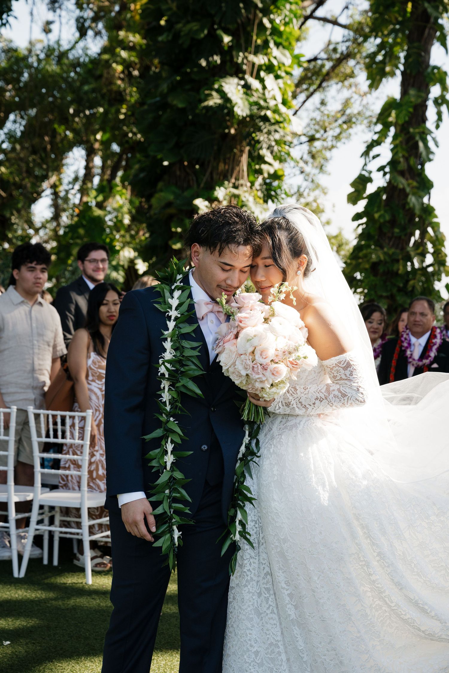 Newlywed couple sharing a tender moment at outdoor wedding ceremony, groom in navy suit with floral lei, bride in lace gown.