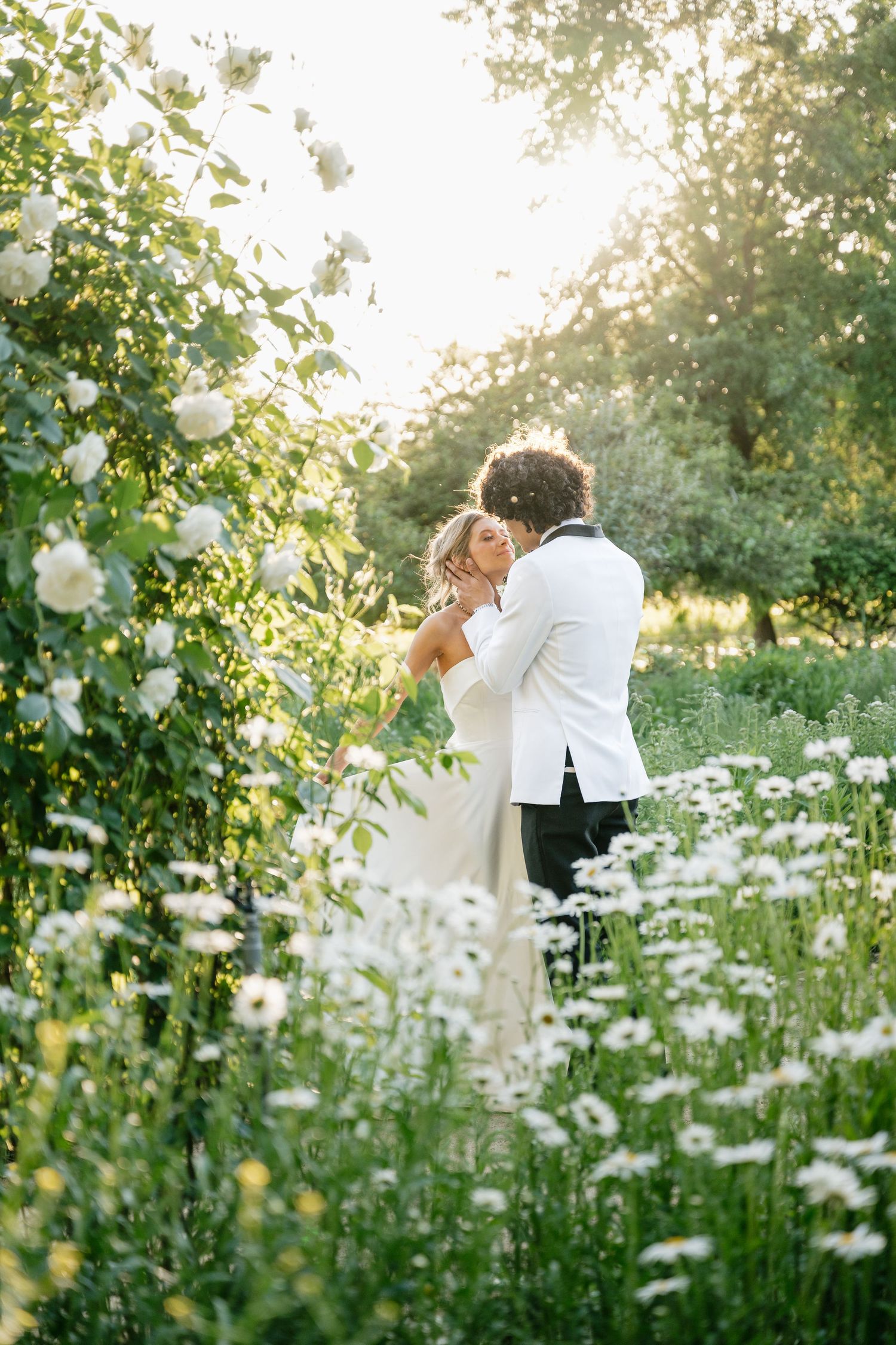 Romantic couple shares an intimate moment in a sunlit garden filled with white wildflowers.