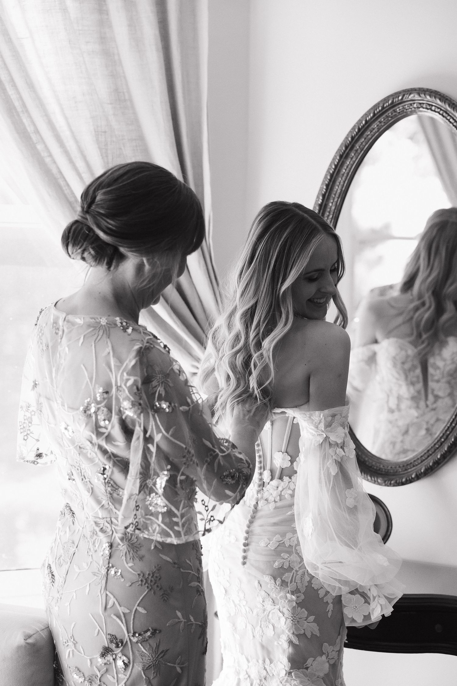 Black and white photo of two women in elegant lace dresses preparing for a wedding by an ornate mirror.