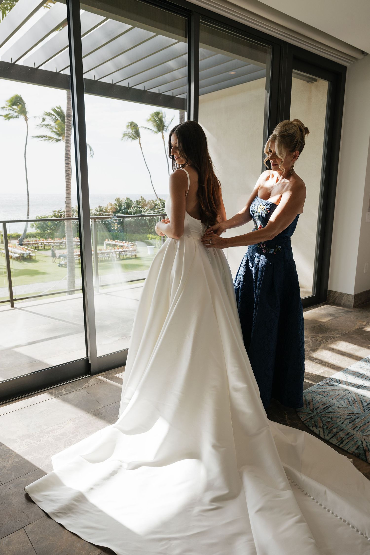 A flowing white wedding dress being adjusted in a bright indoor space with large windows.
