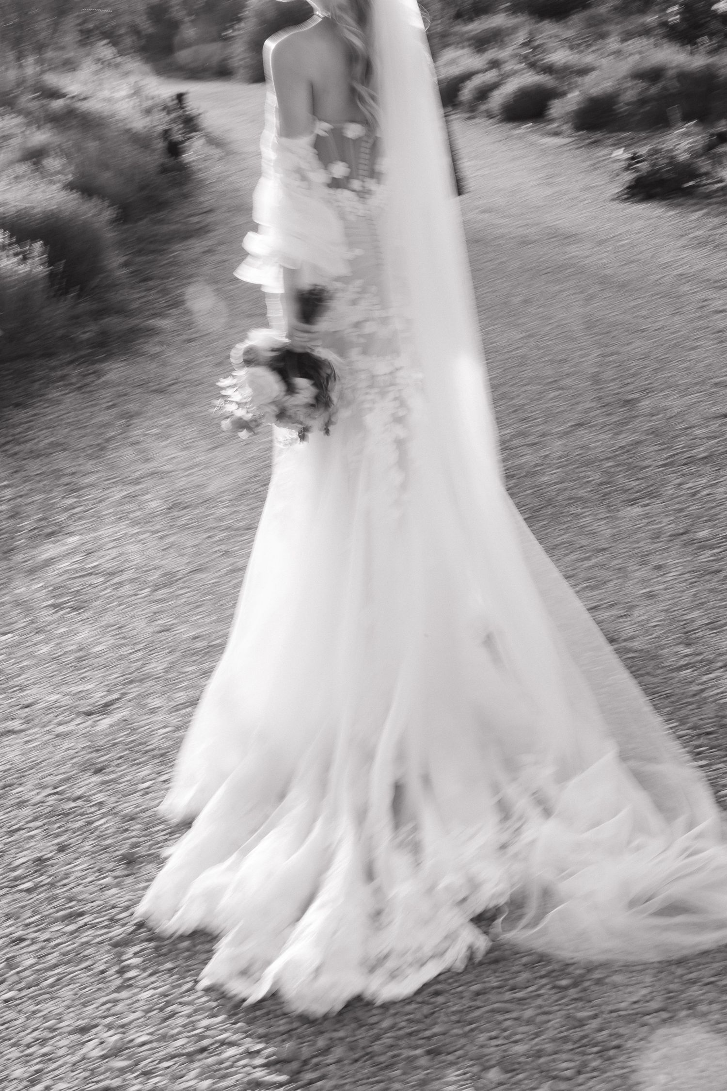 Black and white artistic photo of flowing wedding dress train on garden path.