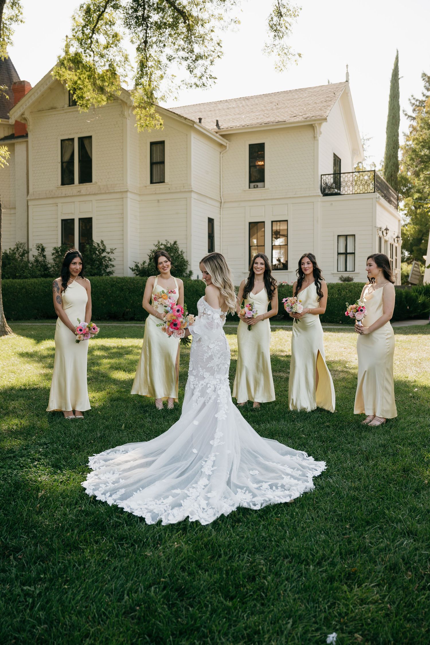 A wedding party poses on a lawn in front of a white two-story house, featuring yellow bridesmaid dresses and a lace train.