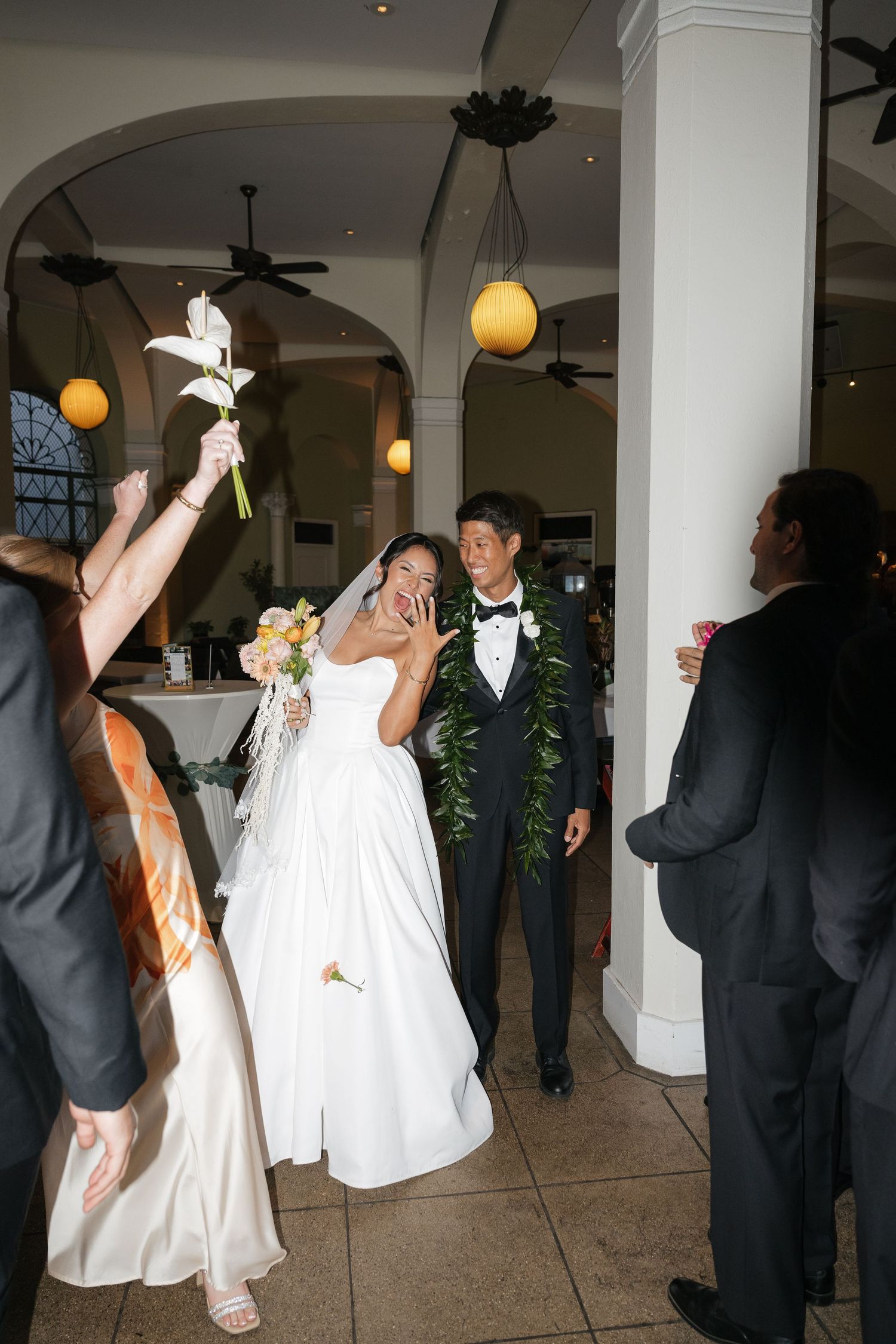 Bride in white gown covers mouth in joy as groom in lei escorts her through venue entrance at Hawaiian wedding.