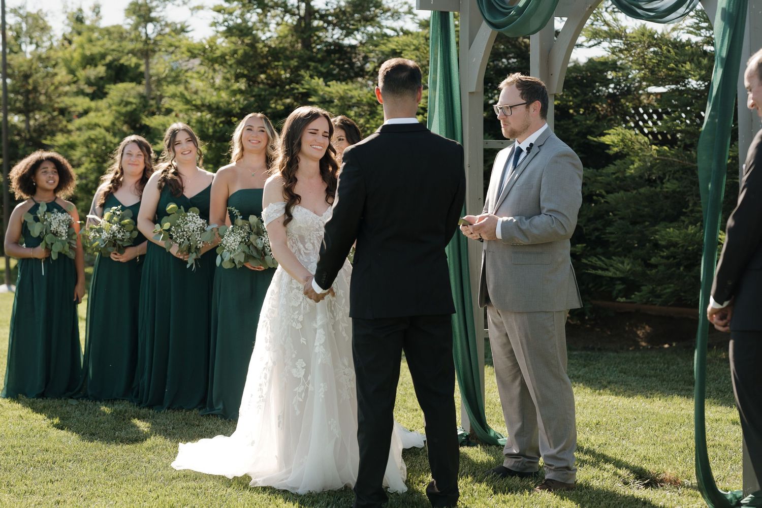 Wedding guests stand in rows on a sunny lawn during an outdoor ceremony with evergreen trees in the background.