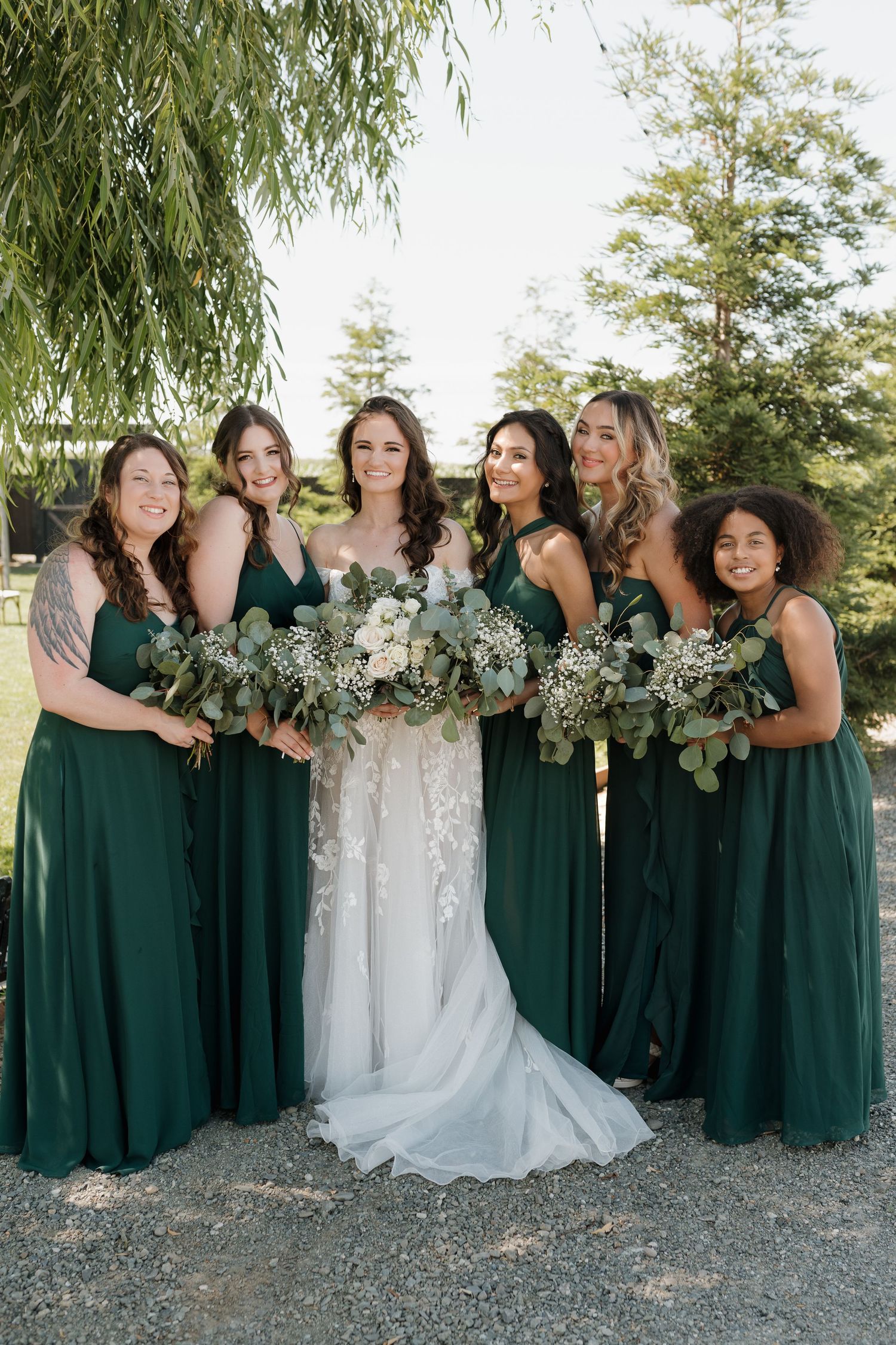 Bridal party in emerald green dresses pose together outdoors under willow trees.