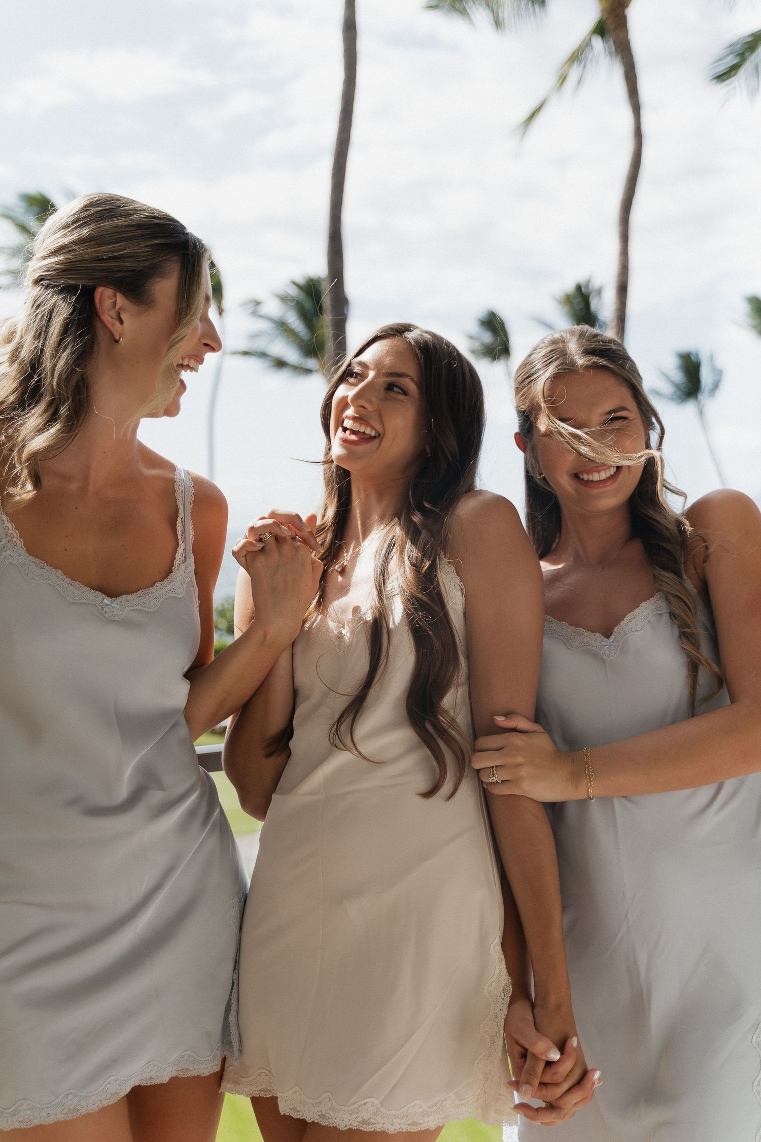 A group of bridesmaids in neutral dresses share joyful laughter together at a tropical beach wedding.