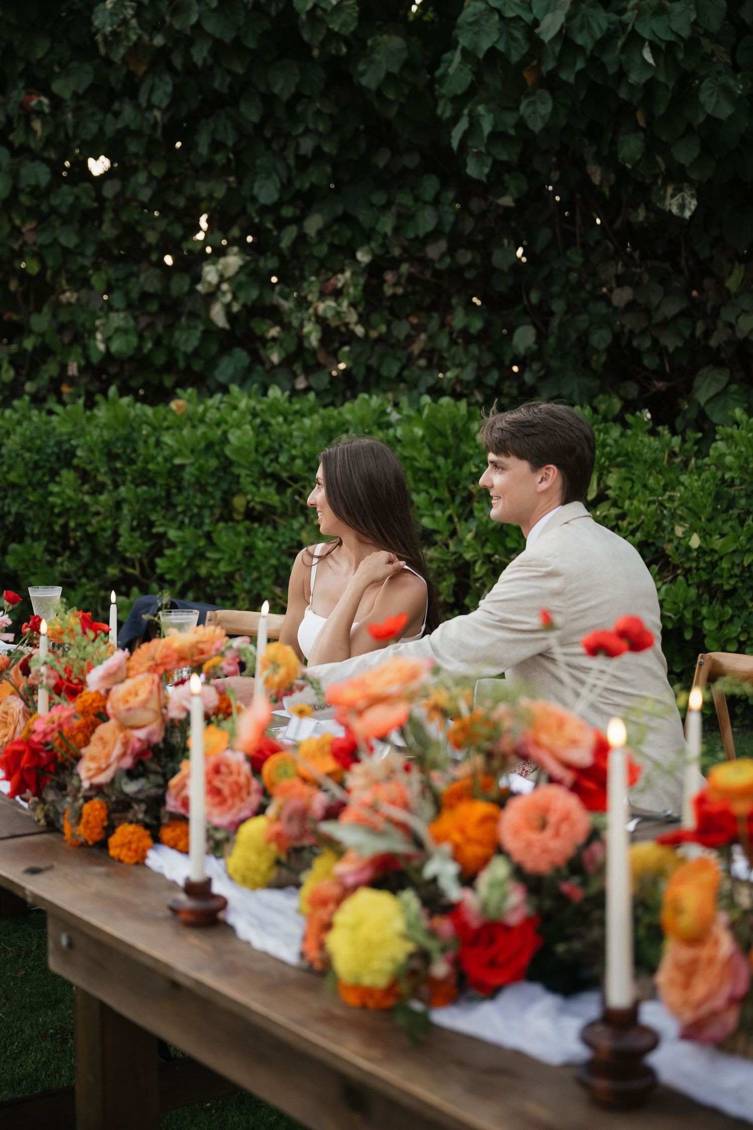 Colorful floral arrangements line a rustic wooden table at an outdoor garden party with orange and yellow blooms.