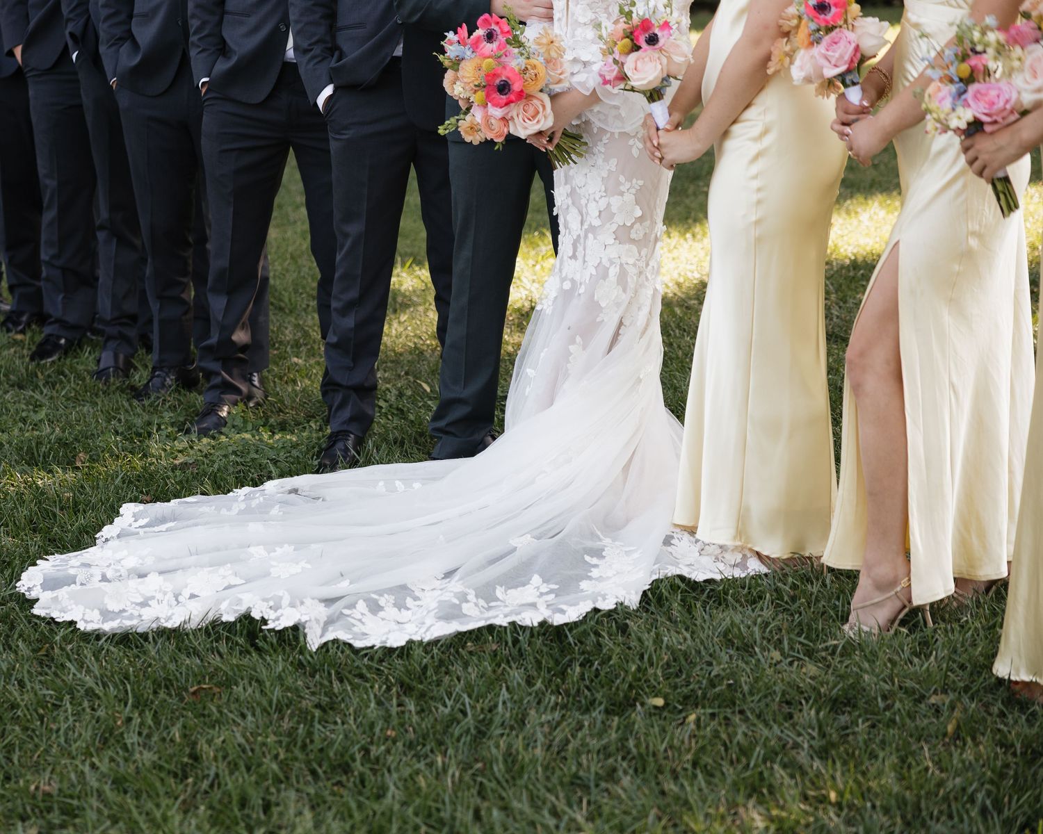 Wedding party members stand together on grass with bridesmaids in yellow dresses and pink bouquets.