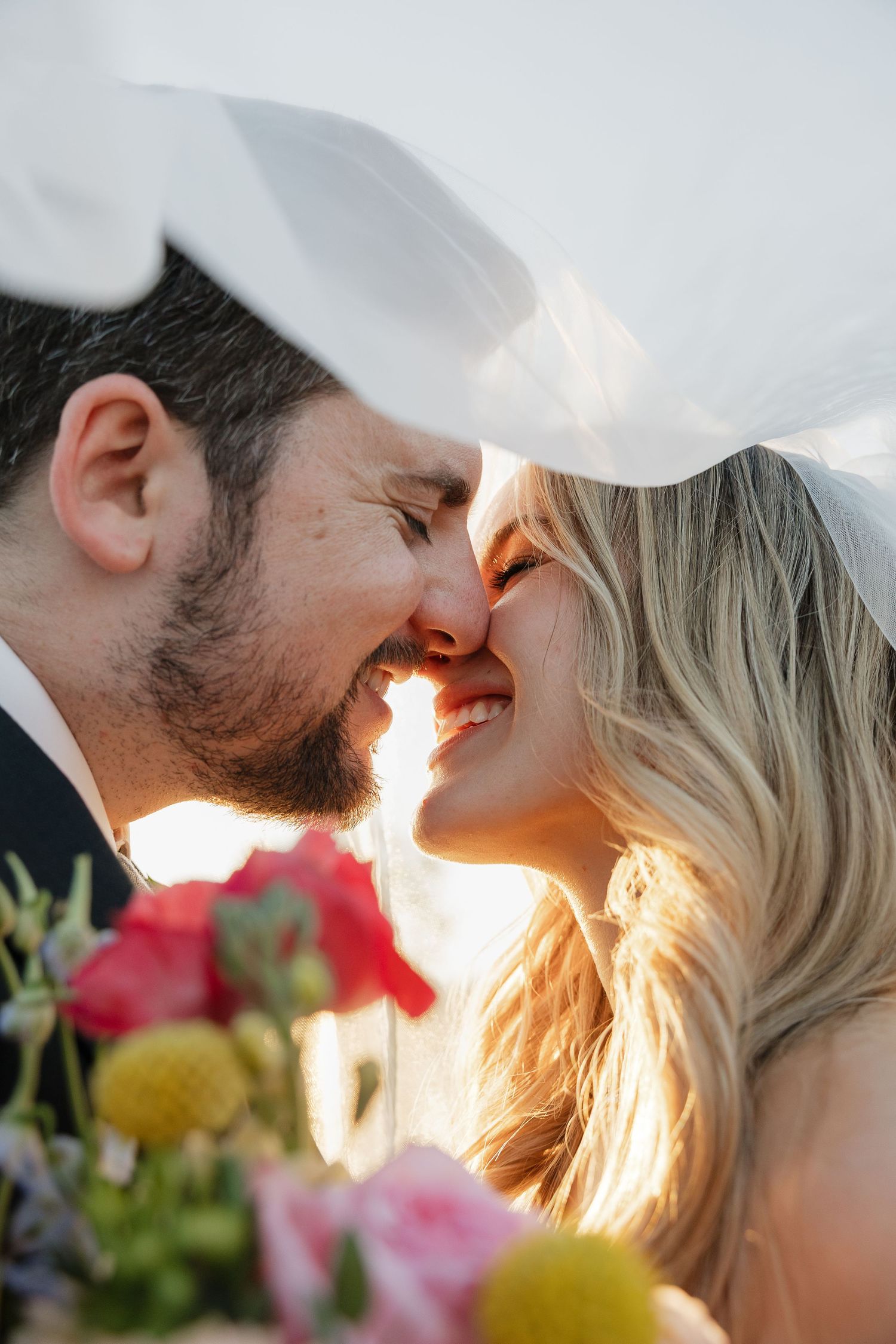 Sunlit close-up of a romantic moment between newlyweds with flowers in the foreground.