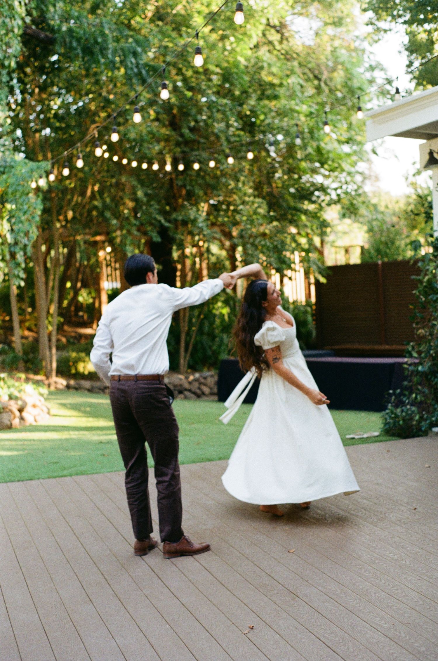 Couple dancing joyfully on a wooden deck outdoors with string lights and greenery creating a romantic atmosphere.