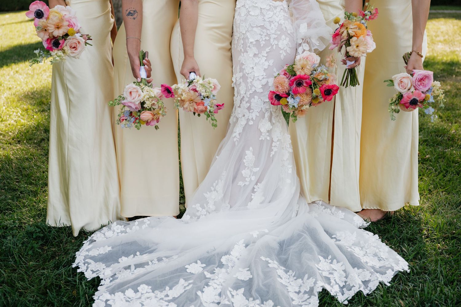 A row of bridesmaids in pale yellow dresses stand next to a bride in an ornate white lace wedding gown with floral bouquets.