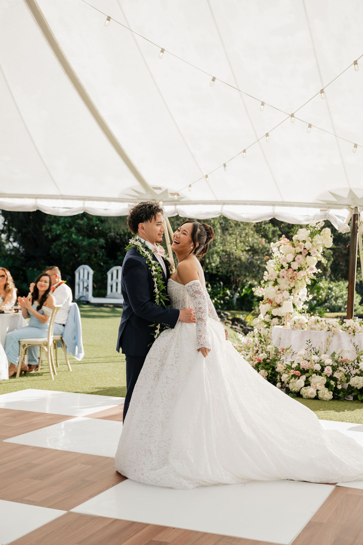 Bride and groom share a joyful first dance under a white tent on a checkered dance floor surrounded by floral arrangements.