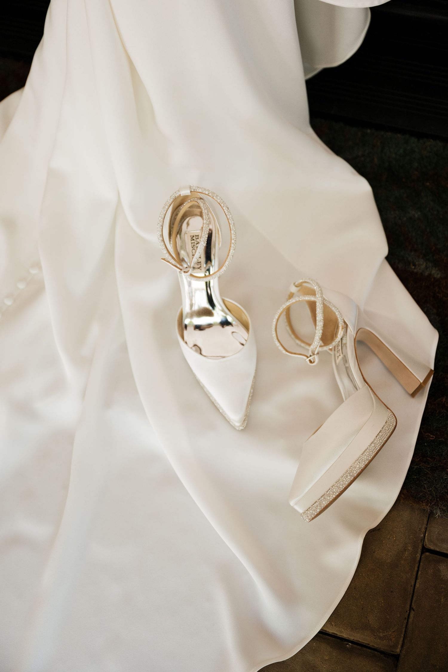 Elegant white bridal heels with gold ankle straps resting on a flowing white wedding dress.