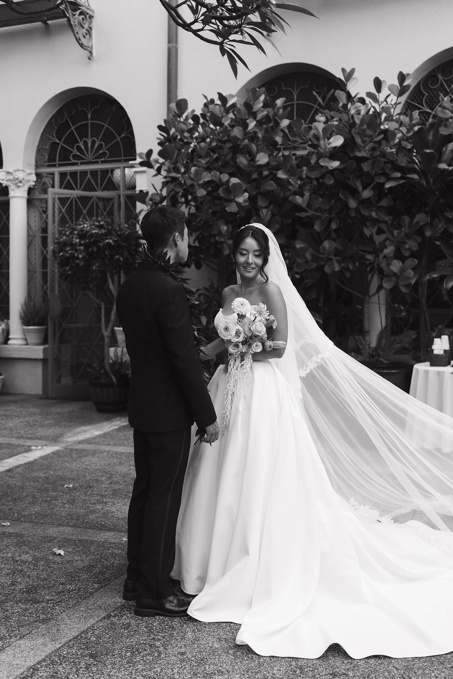 Black and white photo of a smiling bride in a flowing gown and veil holding a bouquet, facing her groom outdoors.
