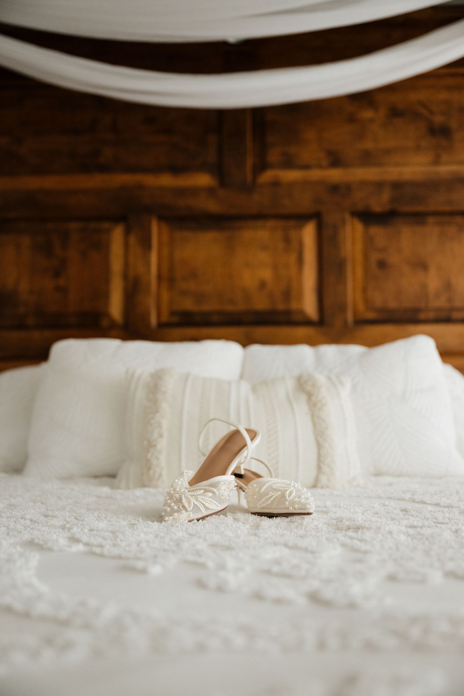 White bridal heels with floral details rest on a white lace bedspread against a wooden headboard.
