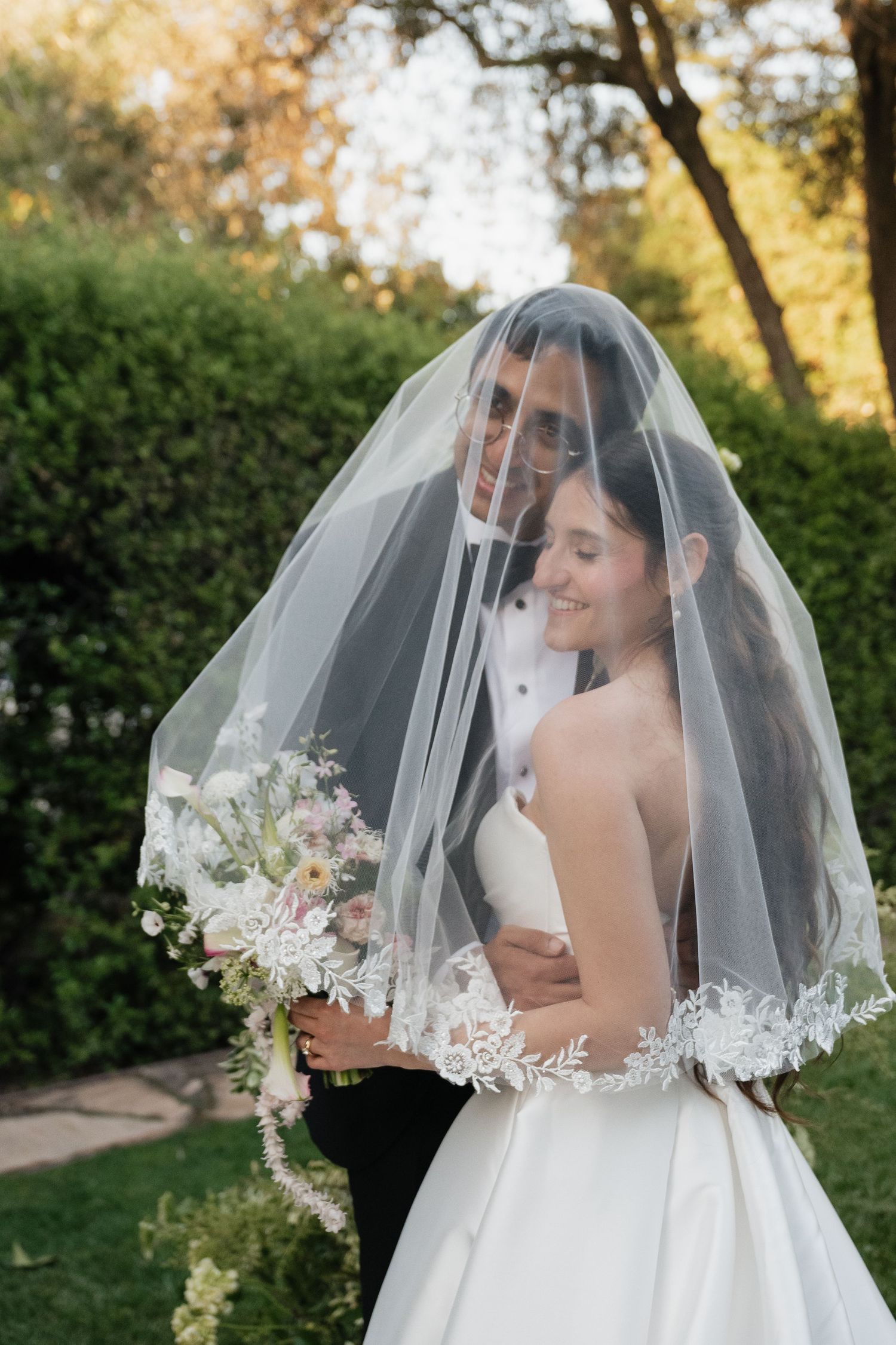 Bride and groom share romantic moment under lace-trimmed veil, bride holding floral bouquet in lush garden setting.