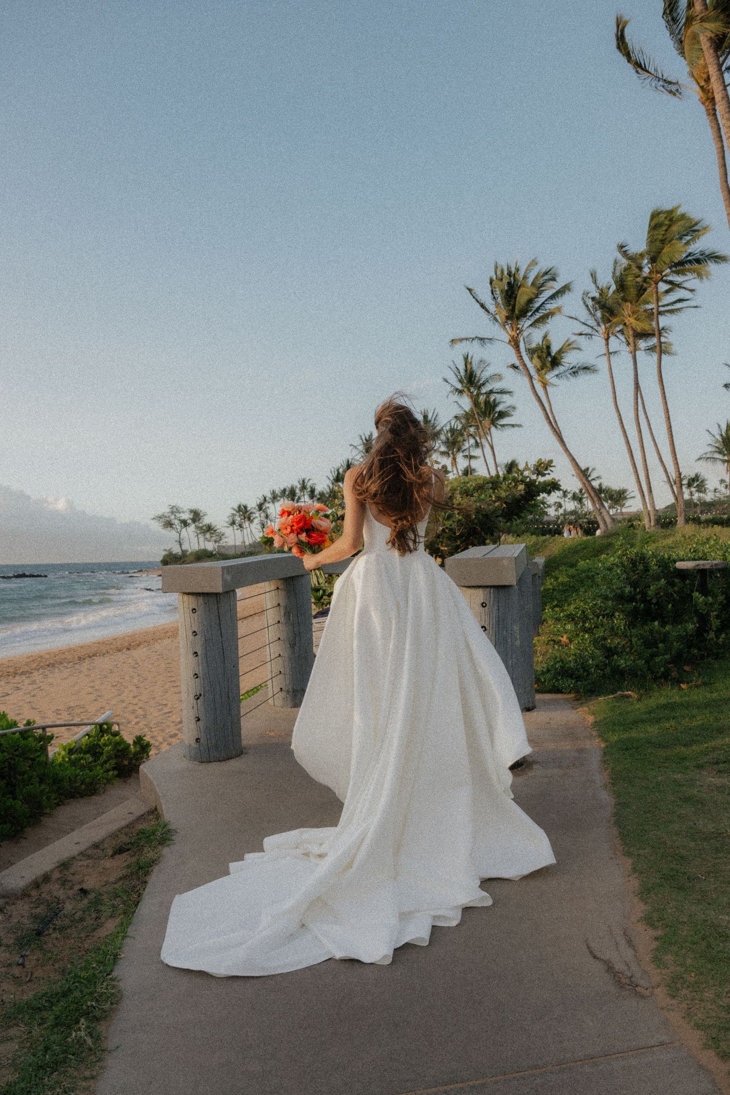 A white wedding gown flows elegantly along a coastal path with palm trees and ocean views in Hawaii.