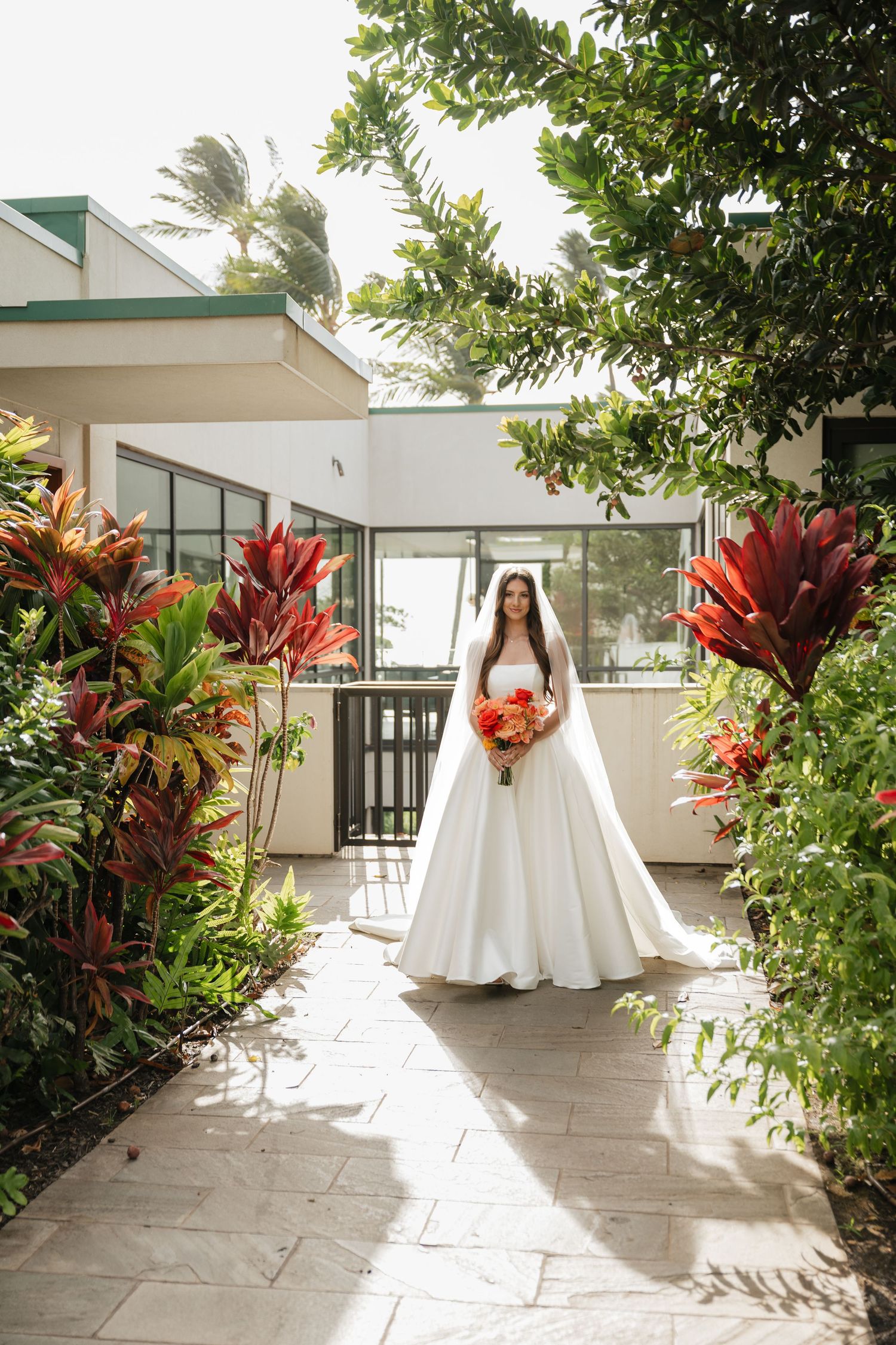 White wedding dress displayed in a tropical garden setting with red flowers and palm trees.