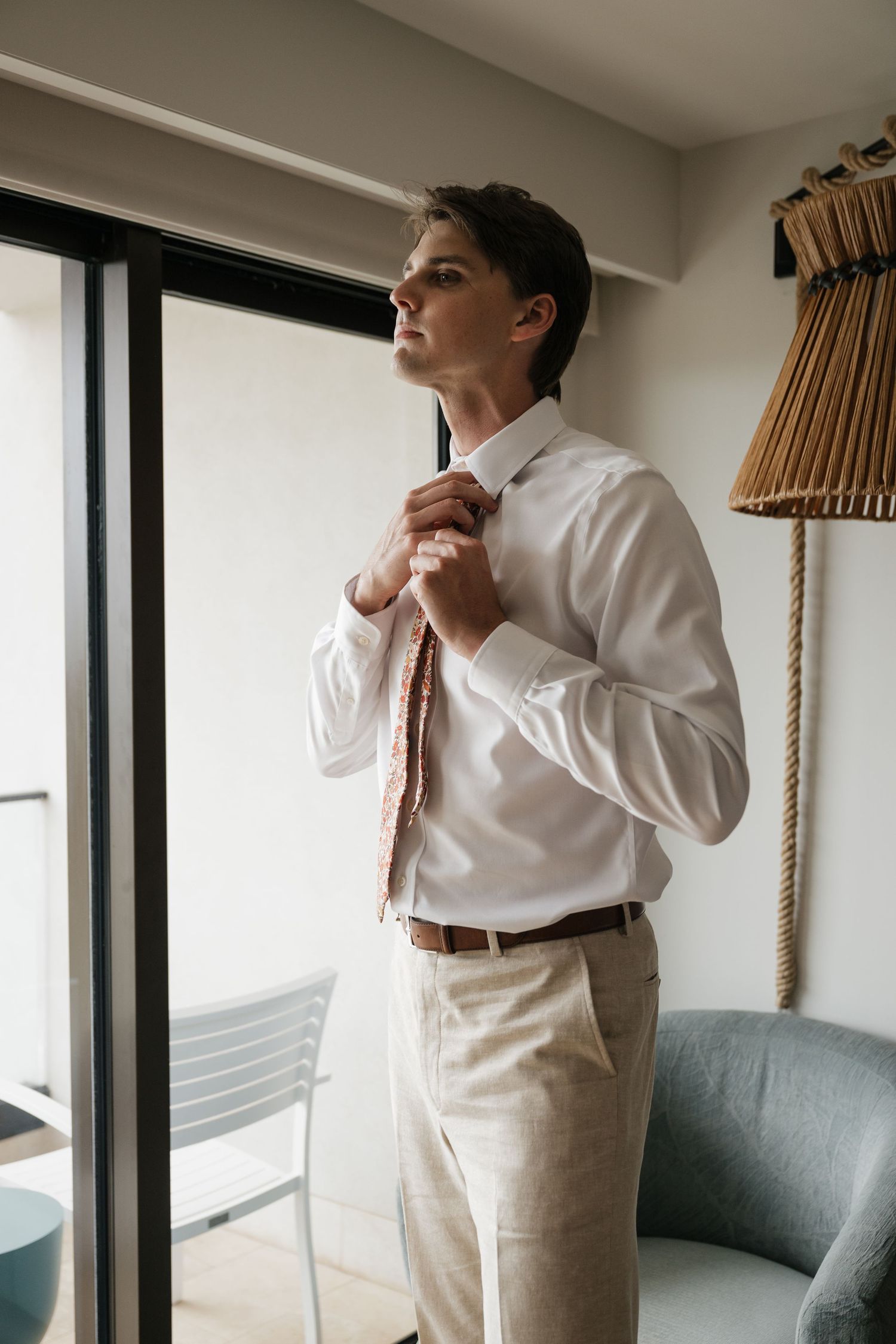 Person adjusting a bow tie while getting dressed in formal attire near a window.