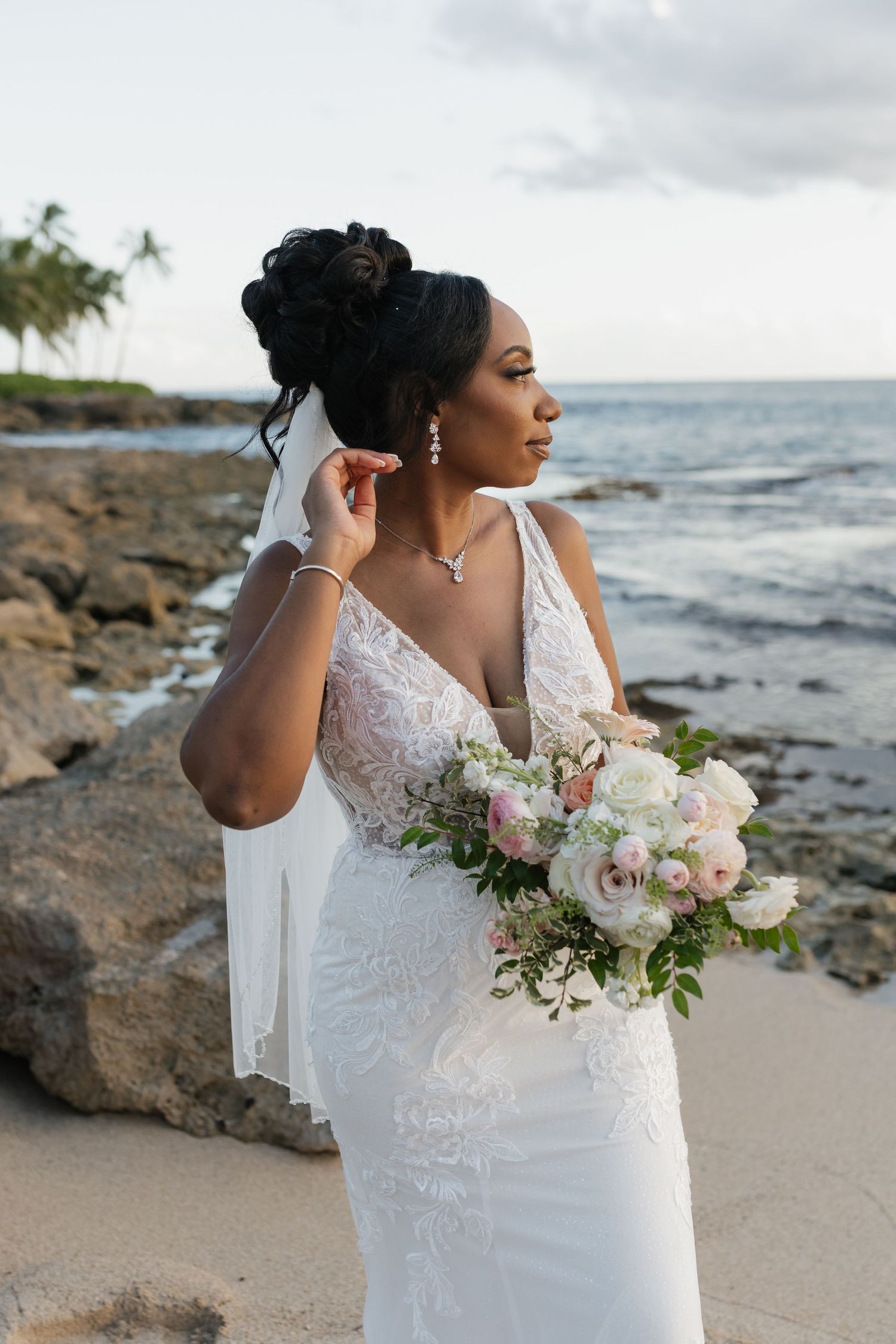 A bride holding a blush and white bouquet poses against an ocean backdrop.