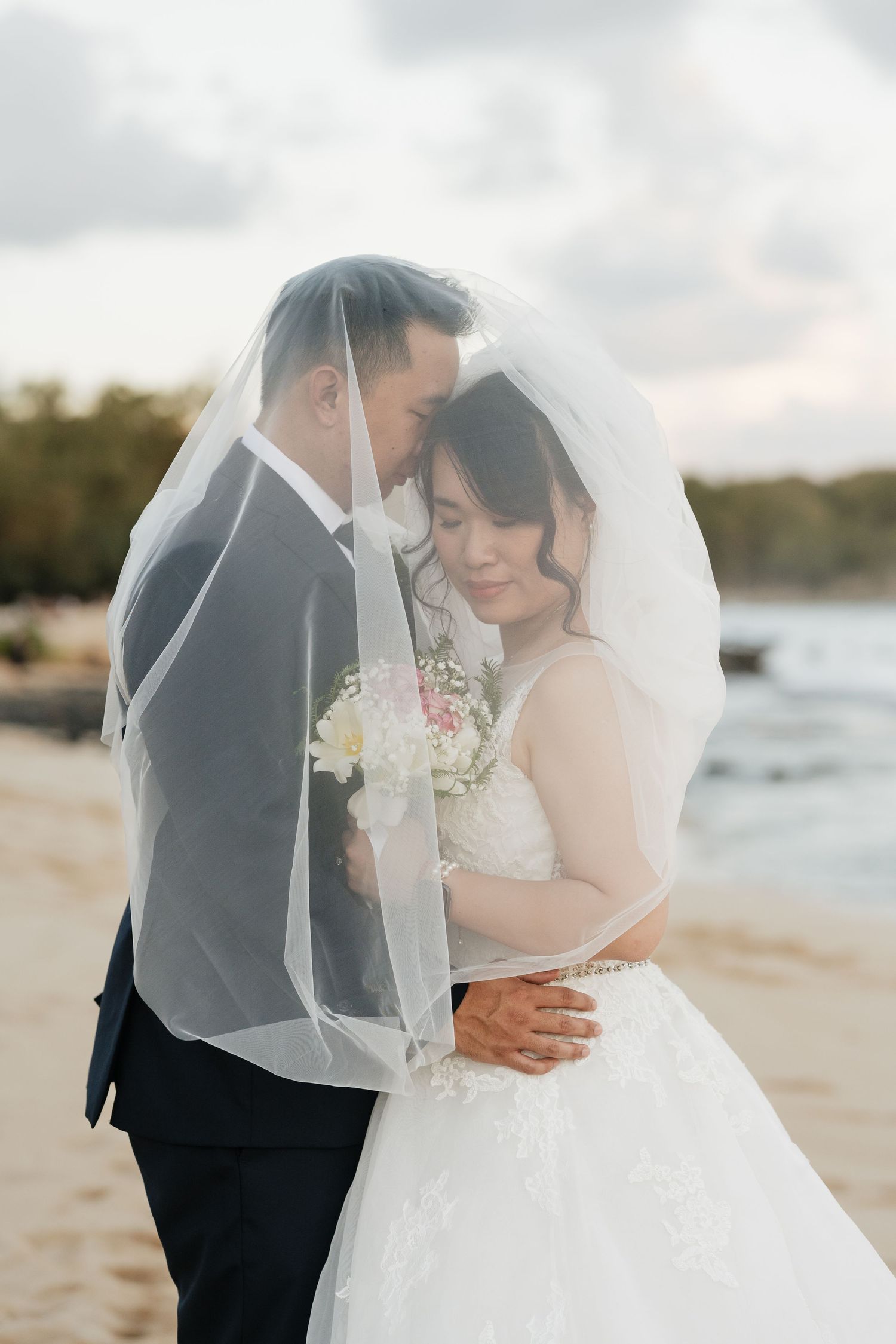 A couple shares an intimate moment under a flowing white veil on a beach at sunset.