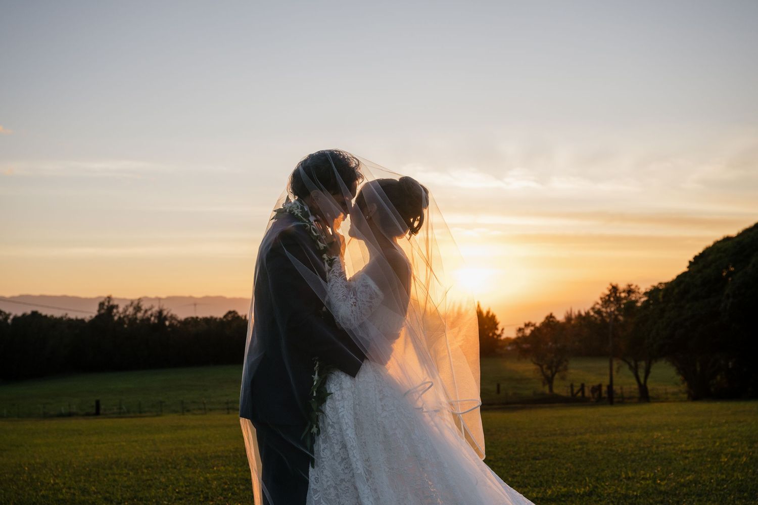 Bride and groom share a romantic kiss at sunset on a green field, with a flowing veil and warm golden sky.