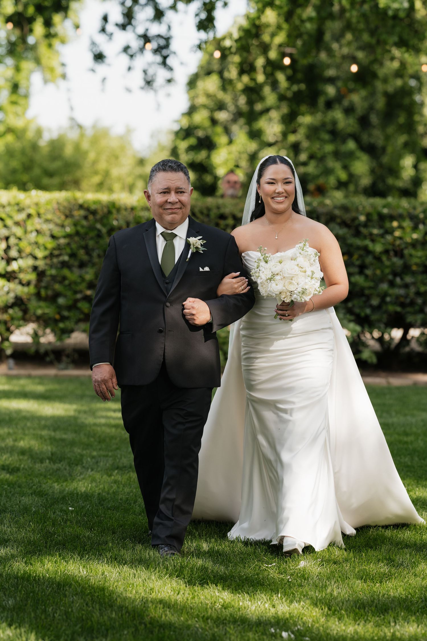 Bride in white gown holding bouquet walks arm-in-arm with suited man across green lawn at outdoor wedding.