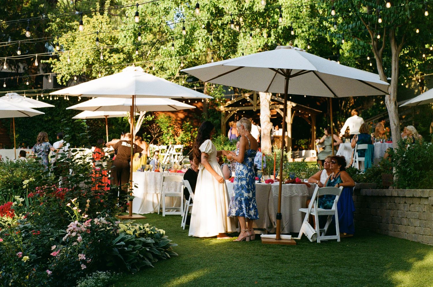 Outdoor garden party with white umbrellas, tables and chairs set up among blooming flowers on a sunny afternoon.